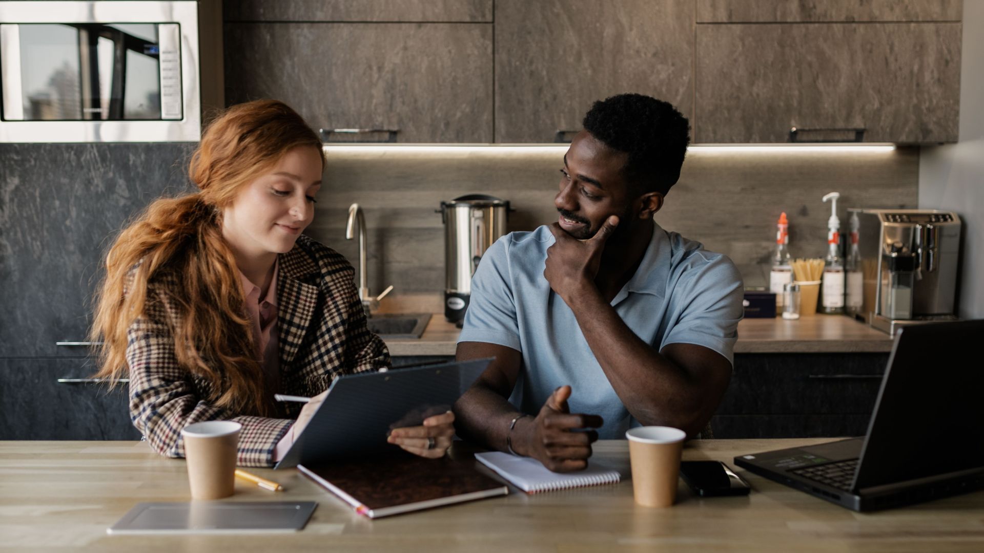 Two colleagues discussing strategies over coffee in a modern office kitchen setting.