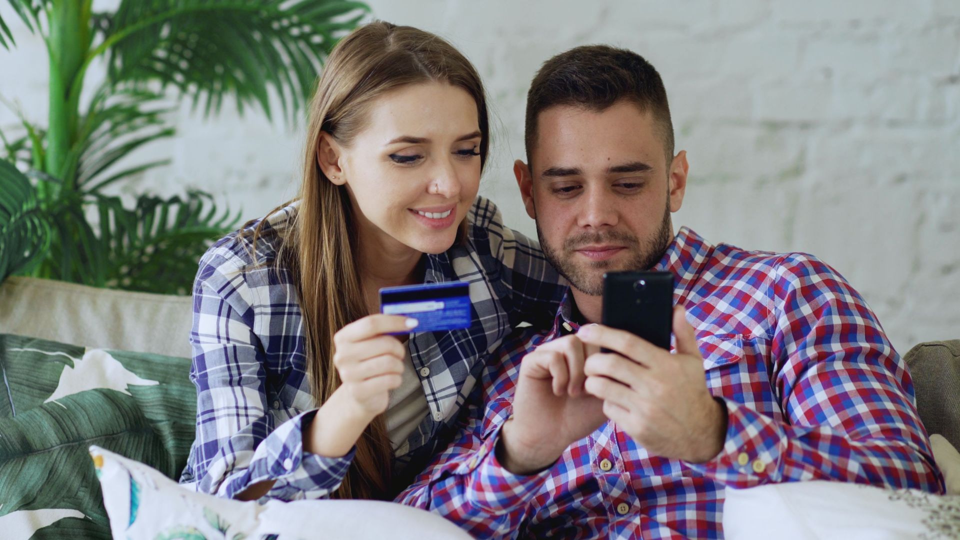 Smiling couple sitting on a sofa, shopping online with a smartphone and credit card.