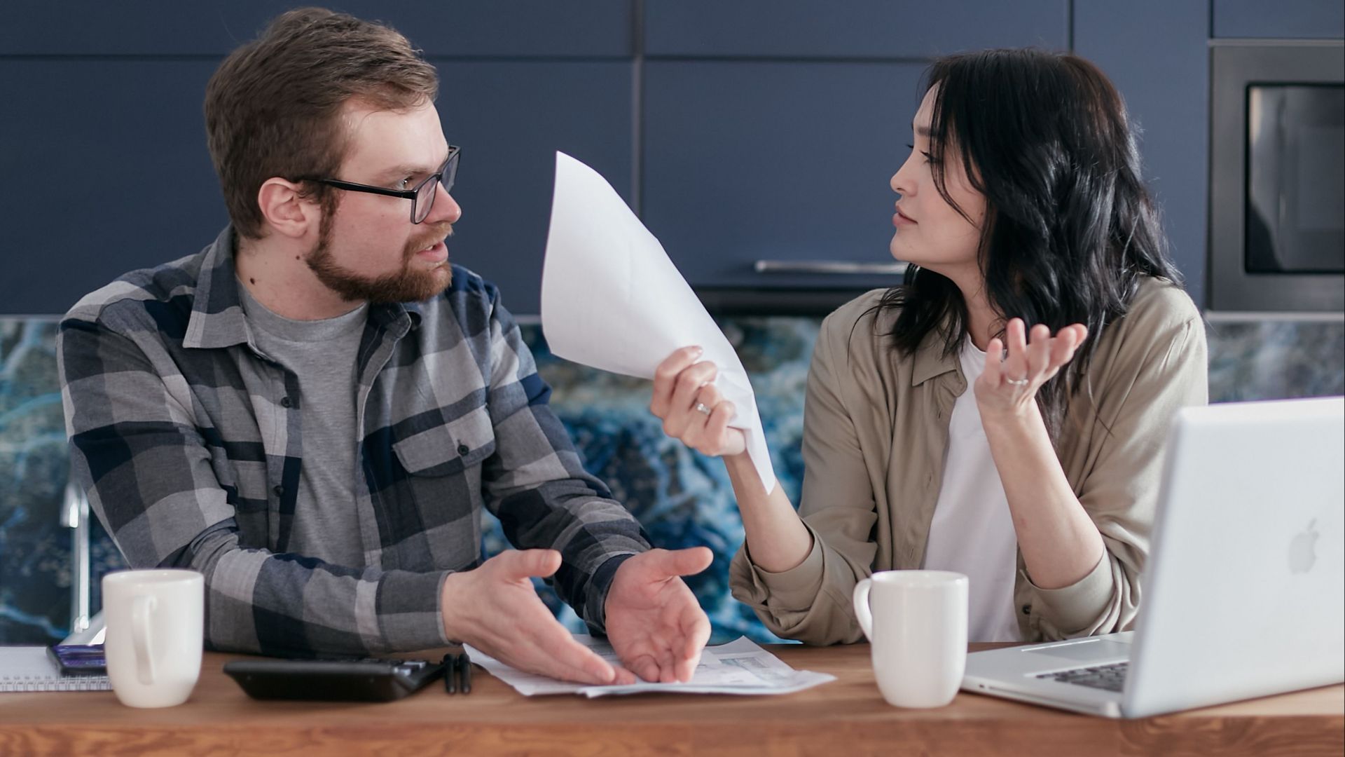 A couple sits at a table, discussing finances with papers and a laptop, looking concerned.