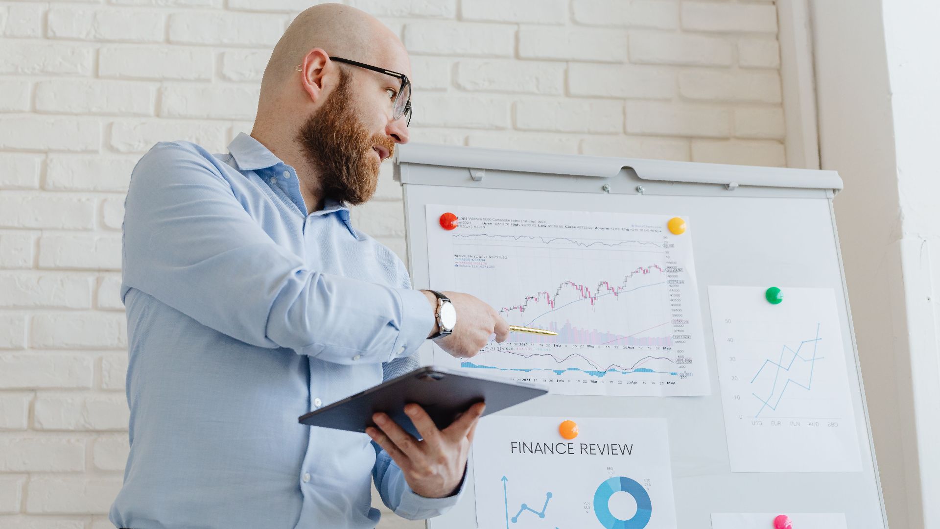 Man giving a business presentation with financial graphs on a whiteboard using a tablet for explanation.