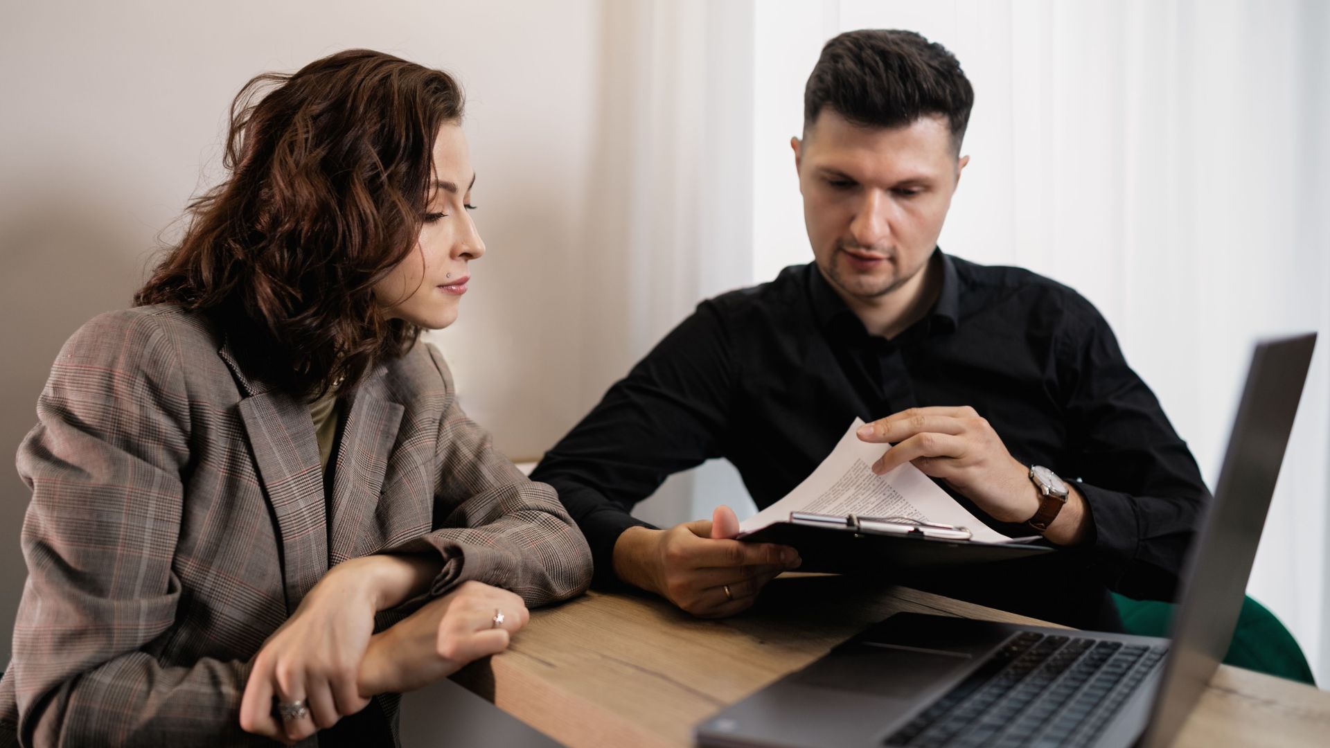 Two professionals discussing documents at a desk with a laptop, focused on collaboration.