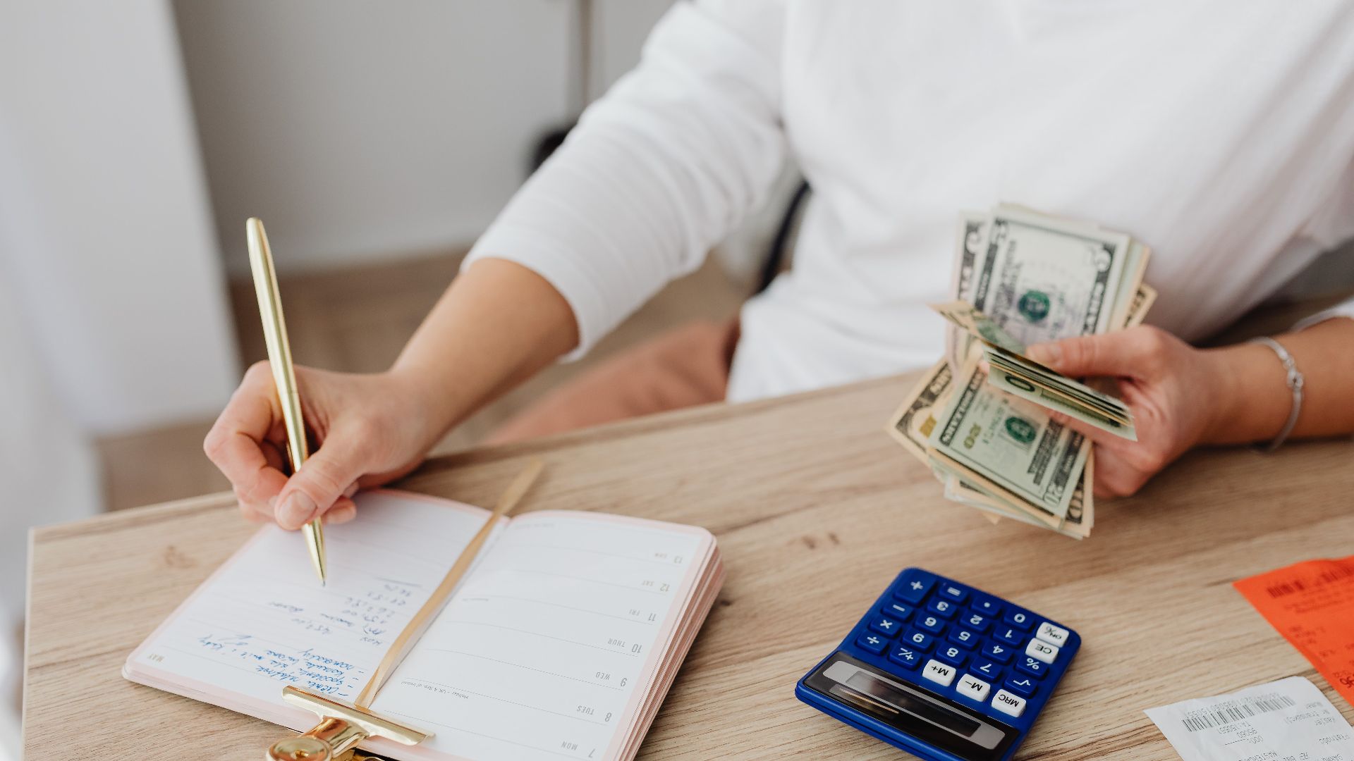 Adult holding cash and writing in planner while using a calculator at home.