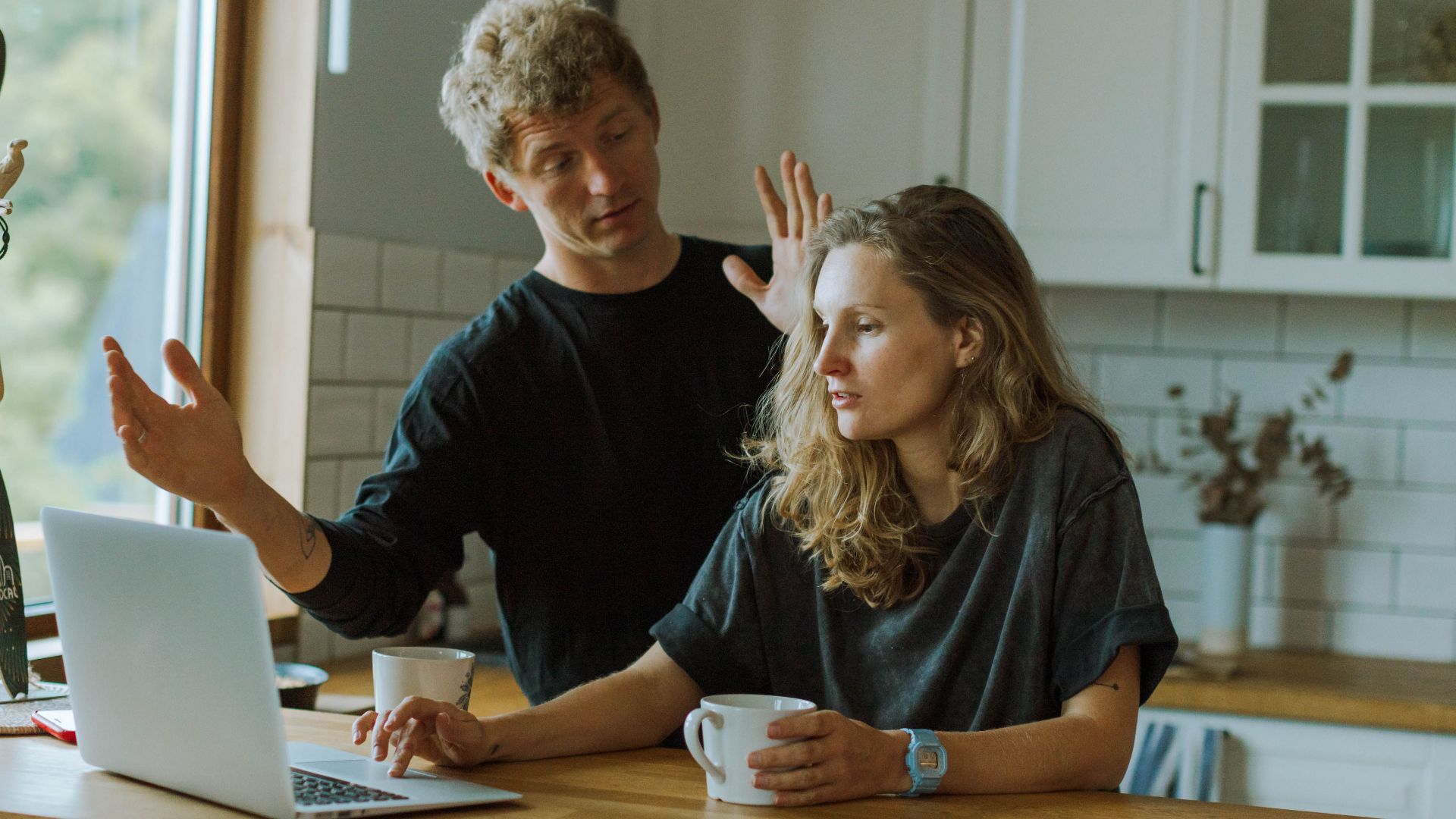 A couple interacting in a bright kitchen using a laptop at a wooden counter.