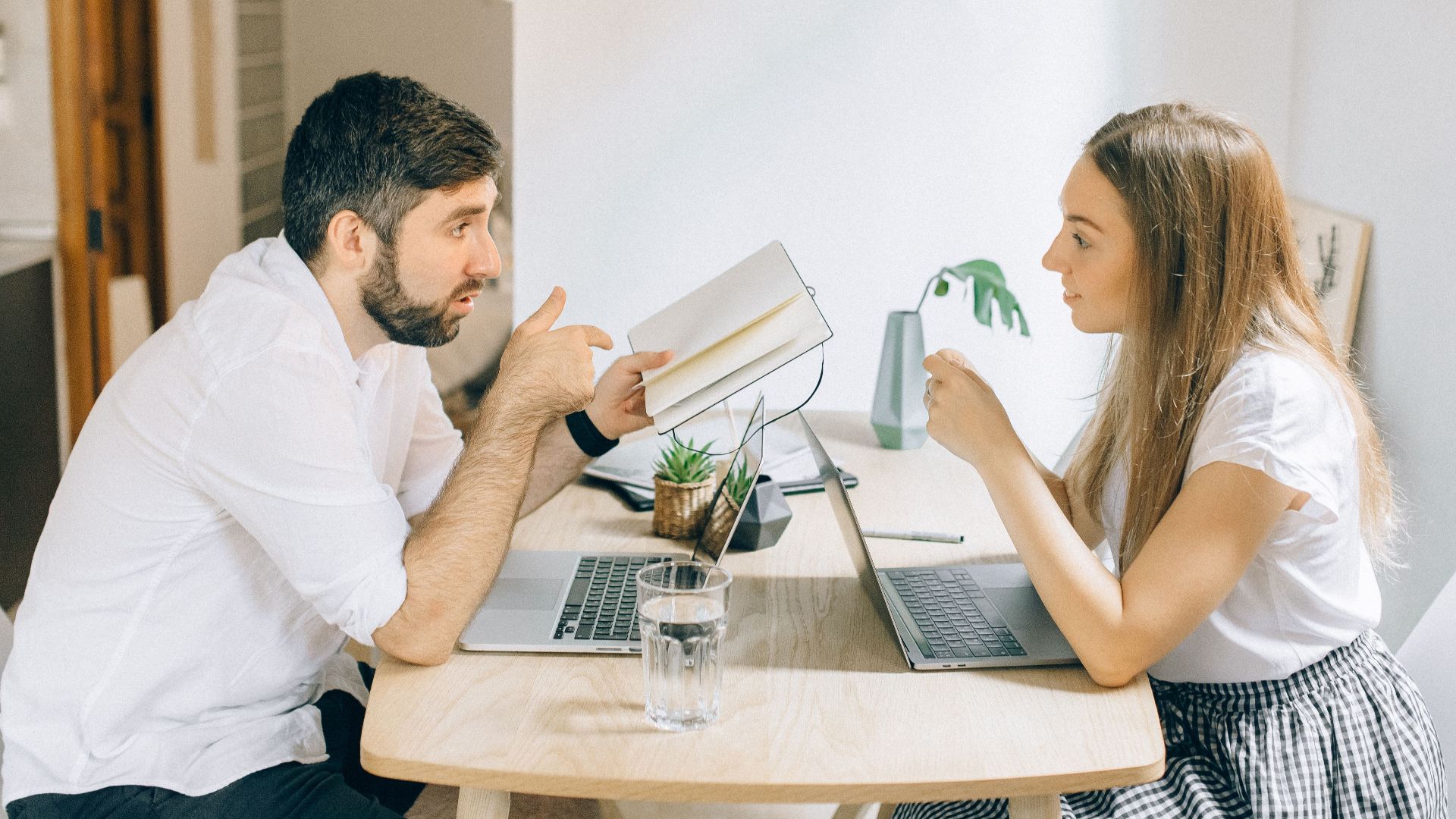 A man and woman working from home on laptops, engaged in discussion at a wooden table.