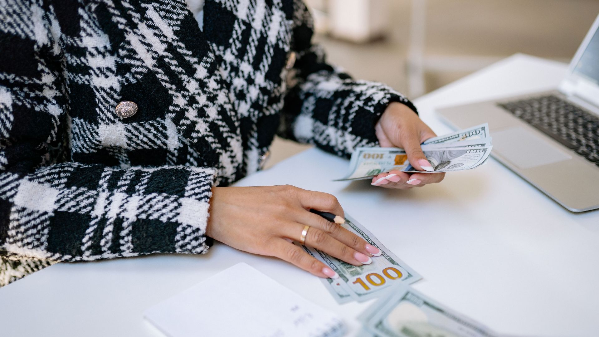 Close-up of a woman counting hundred-dollar bills at a desk with a laptop, focusing on finance and work.