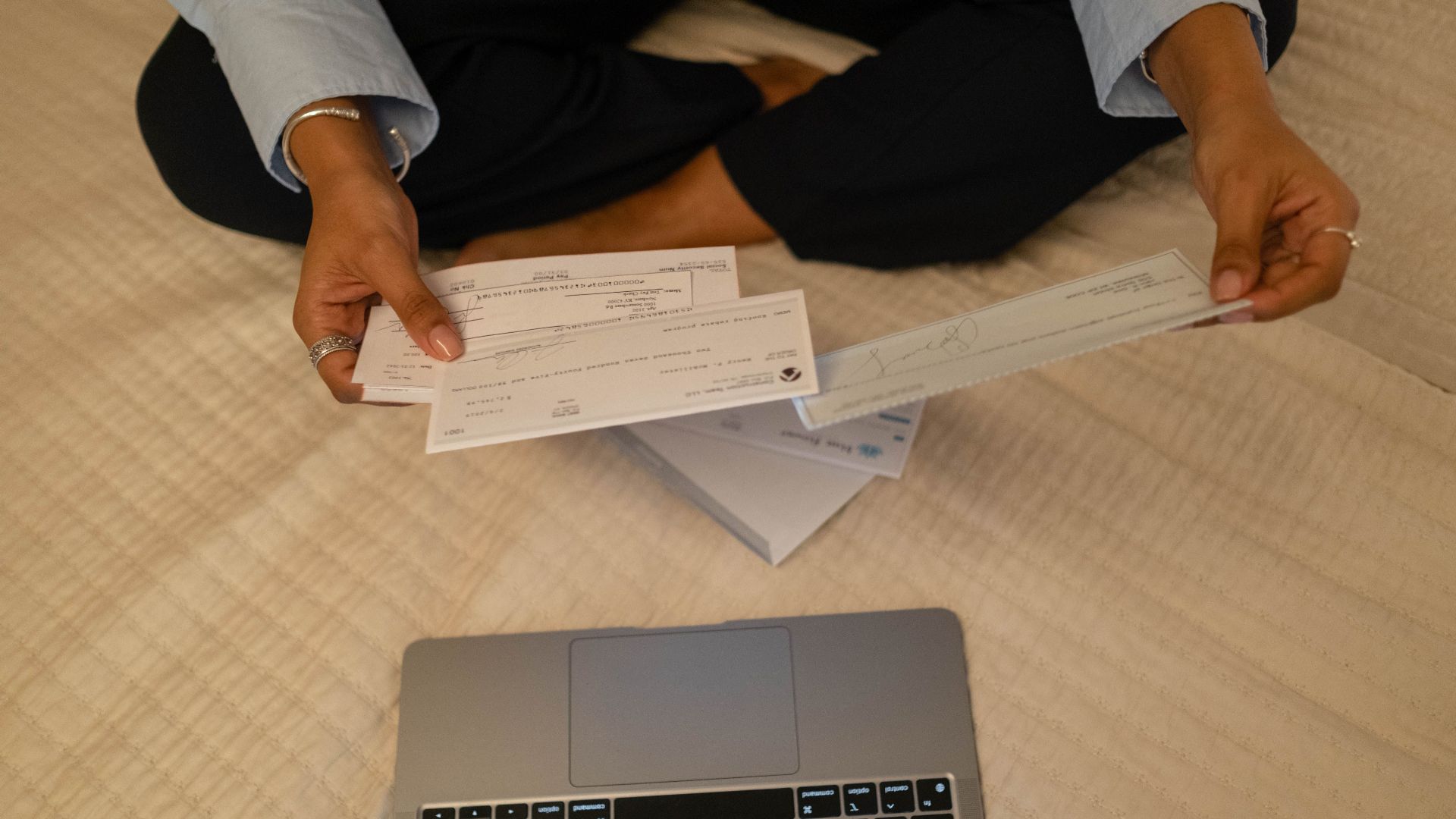 Close-up of hands holding cheques beside a laptop indoors for financial tasks.