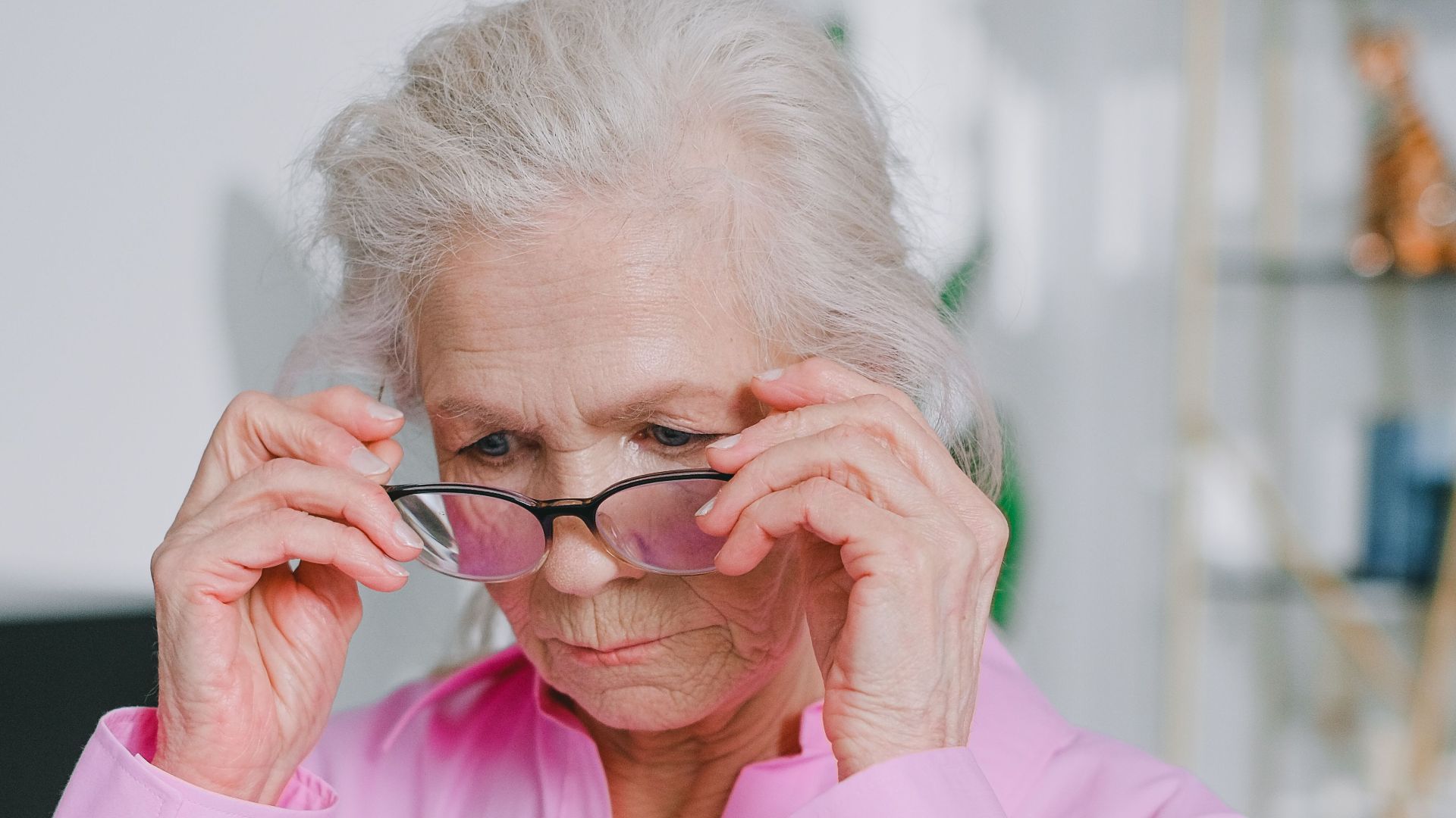 Senior woman adjusting eyeglasses while reading a document indoors, wearing a pink shirt.