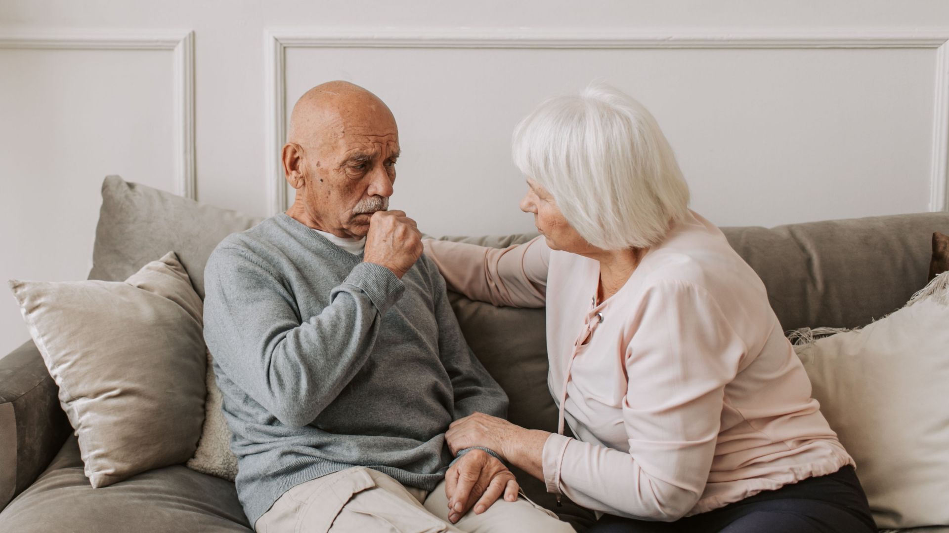 An elderly woman comforts a man coughing on a couch, showcasing care and affection.