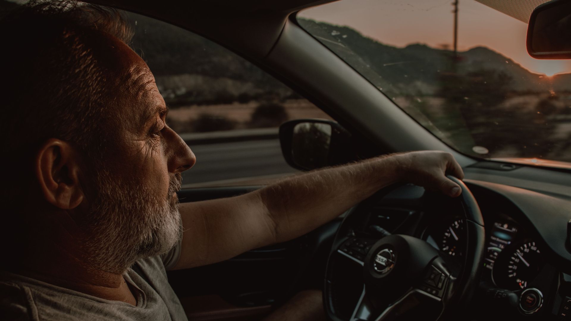 Elderly man driving a Nissan car during sunset, depicting travel and contemplation.