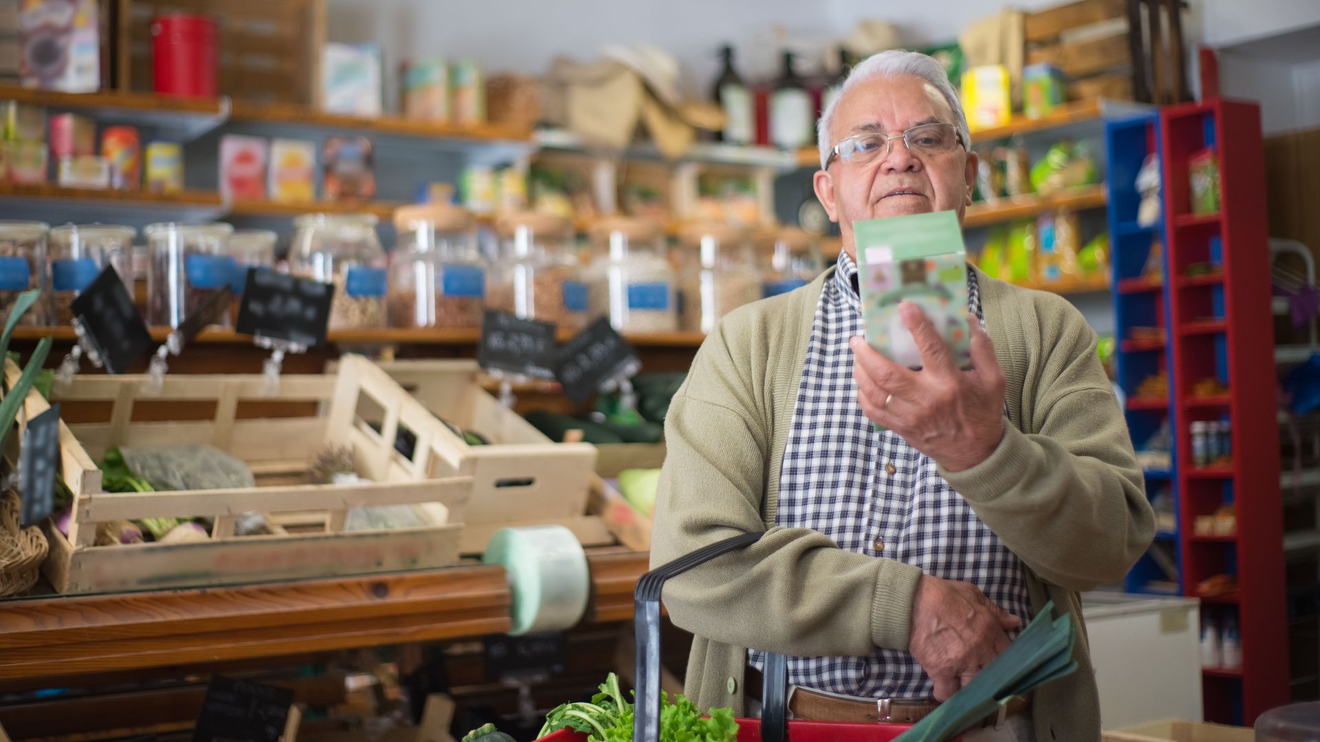 Elderly man examines product in grocery store aisle, representing daily shopping routine.