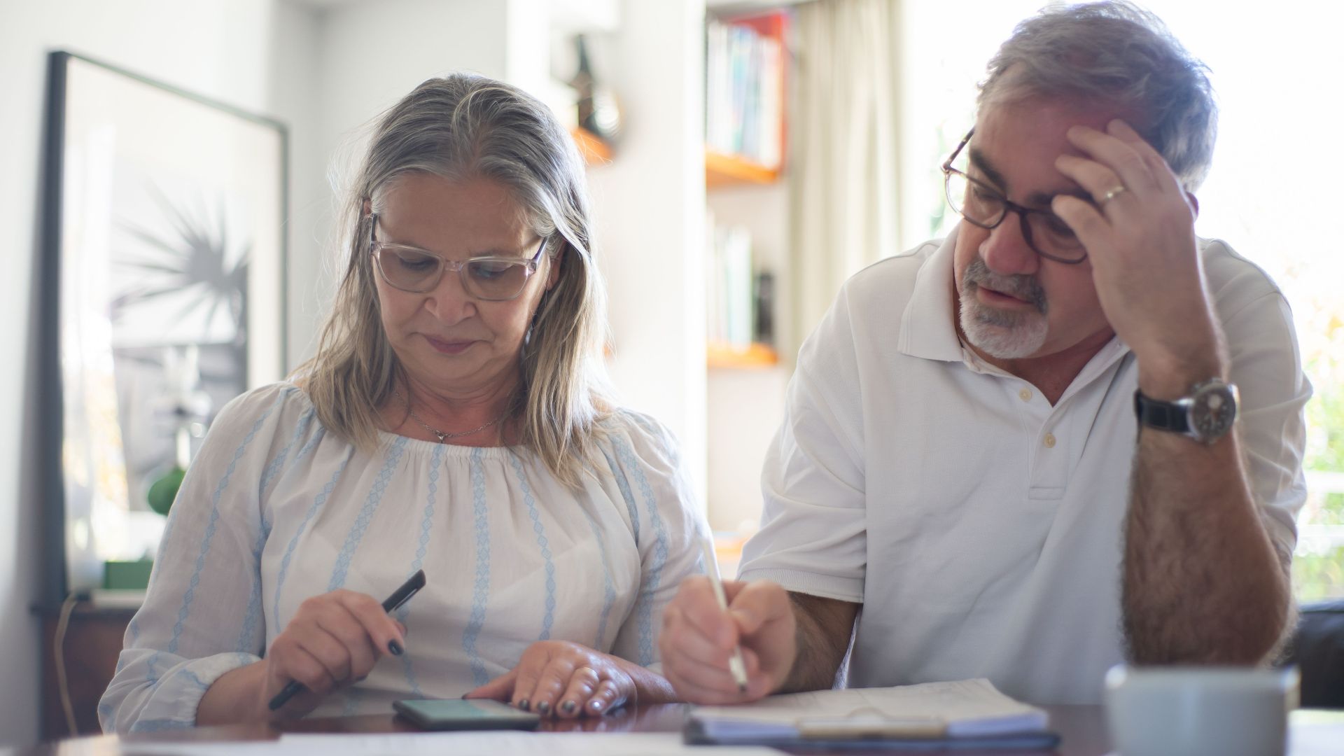 Senior couple calculating expenses at home office desk with documents and notes.