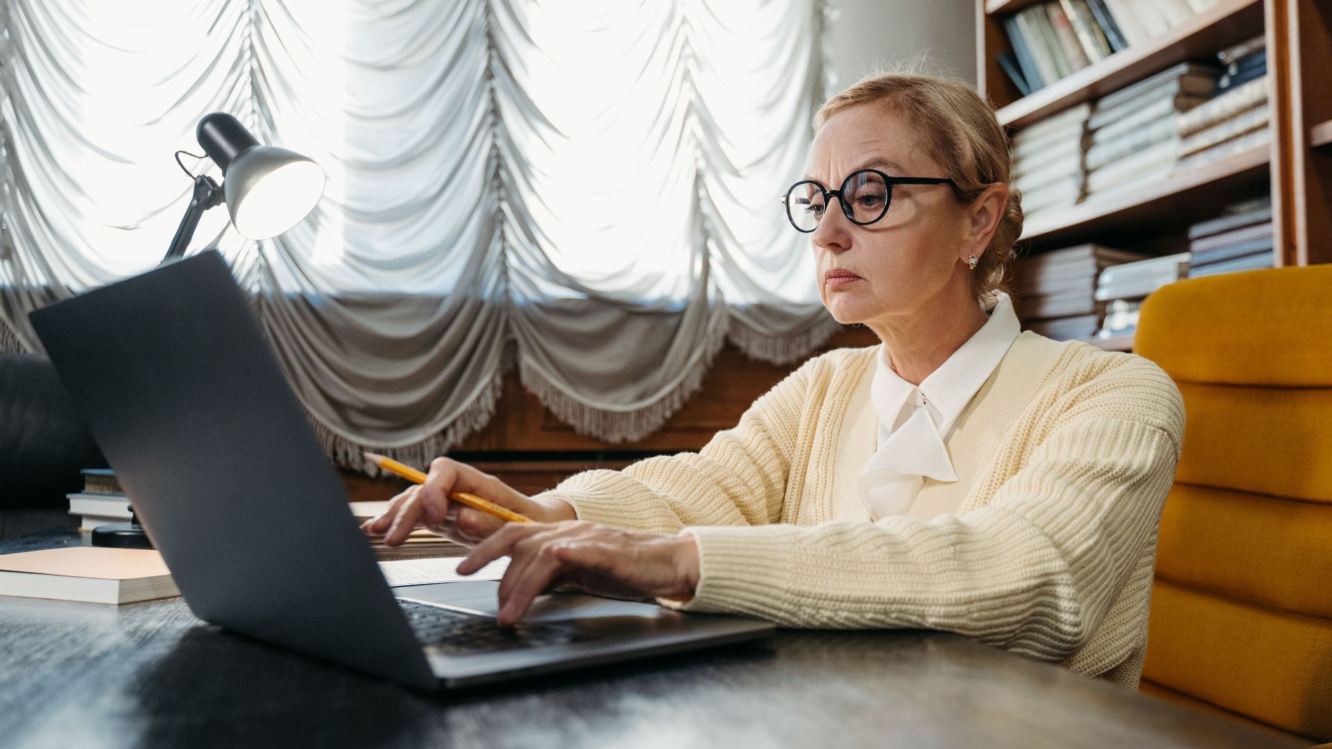 Mature woman in glasses working intently on a laptop at a library desk.