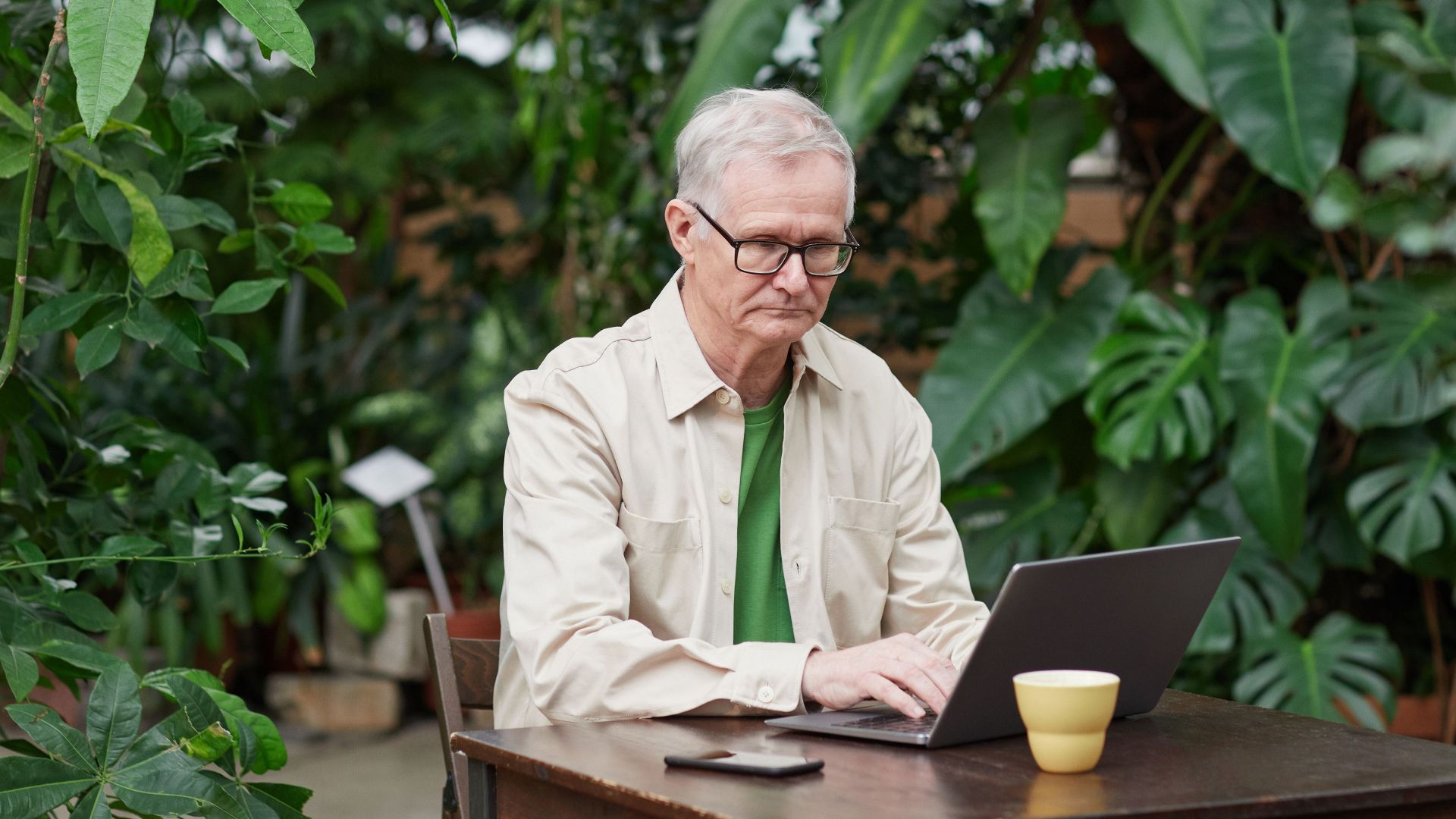 Elder man using laptop in a lush outdoor setting, showcasing freelance remote work lifestyle.