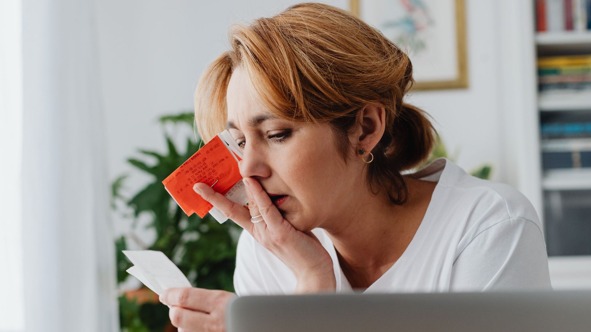 Woman concerned over finances with laptop, card, and receipts. Indoor, modern home office setting.
