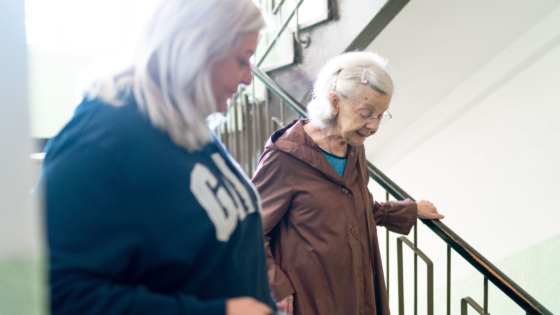 Elderly woman with caregiver on stairs, symbolizing home care and support, in Prague setting.