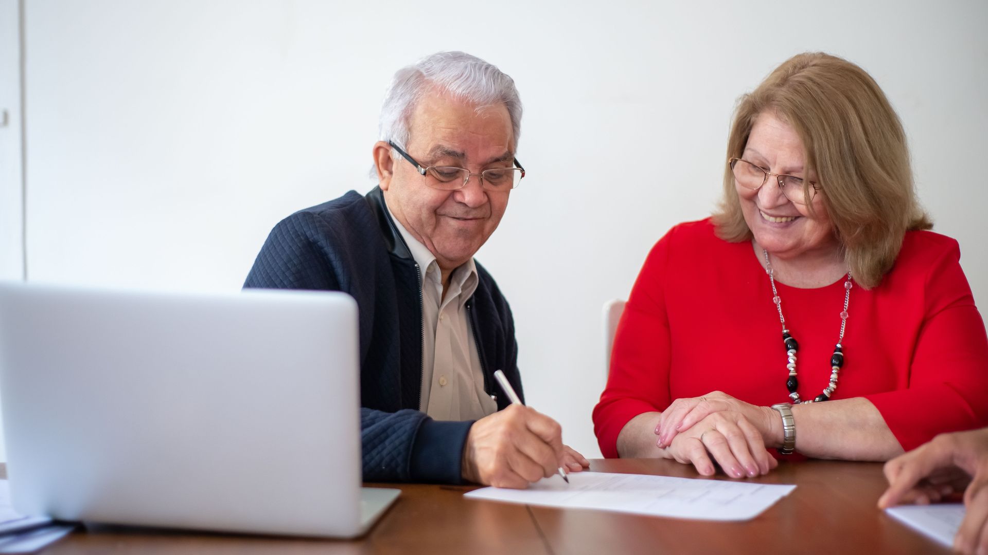 Elderly couple signing documents at a desk with a laptop. Smiling and working.