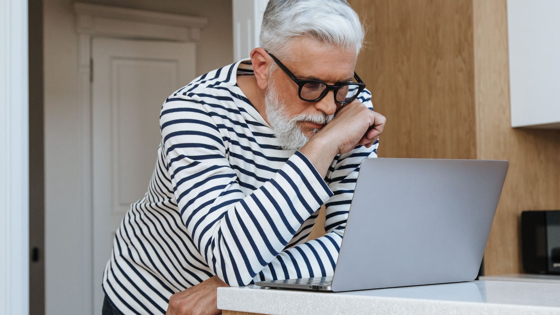 A senior adult man with glasses concentrating on his laptop at home, symbolizing modern working environments.