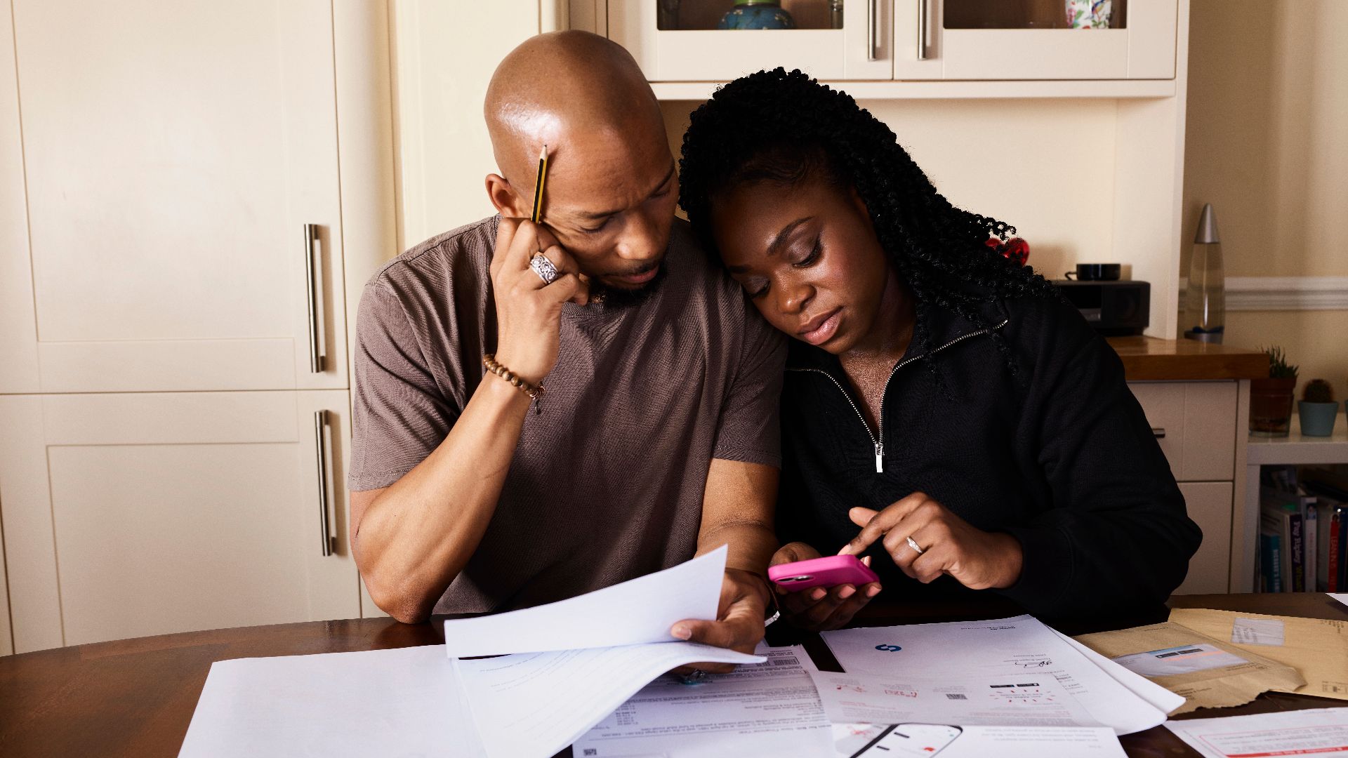 A couple sits at a table managing domestic finances, evaluating documents and using a smartphone.