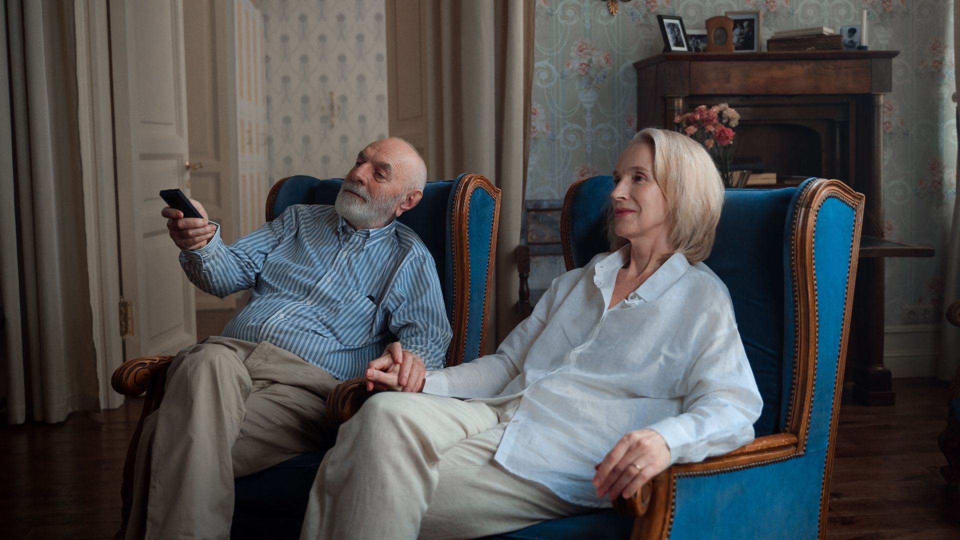 An elderly couple sits holding hands, enjoying a peaceful moment at home.