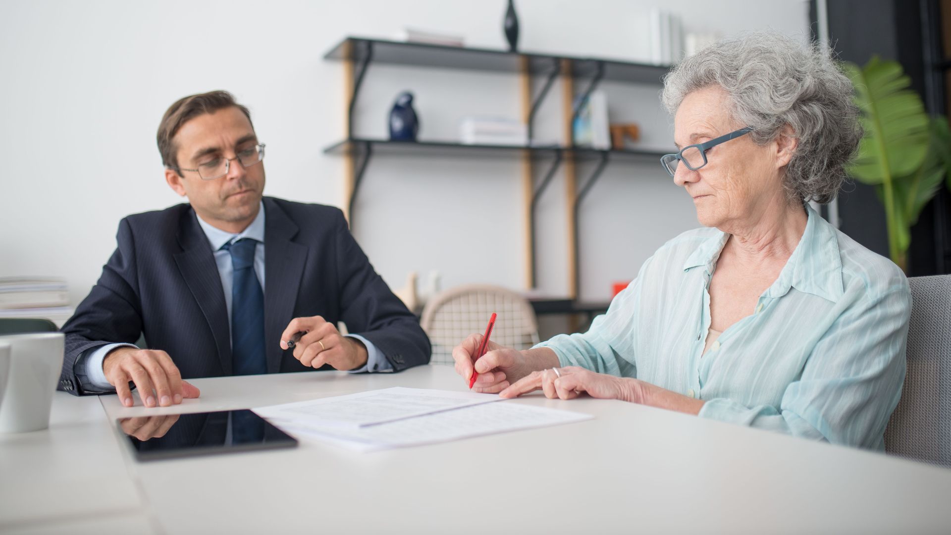 Elderly woman signing paperwork in modern office with consultant.