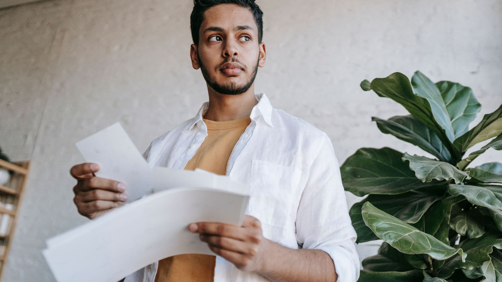 Thoughtful ethnic male in casual clothes holding printed pictures in hands and looking away while standing in light living room