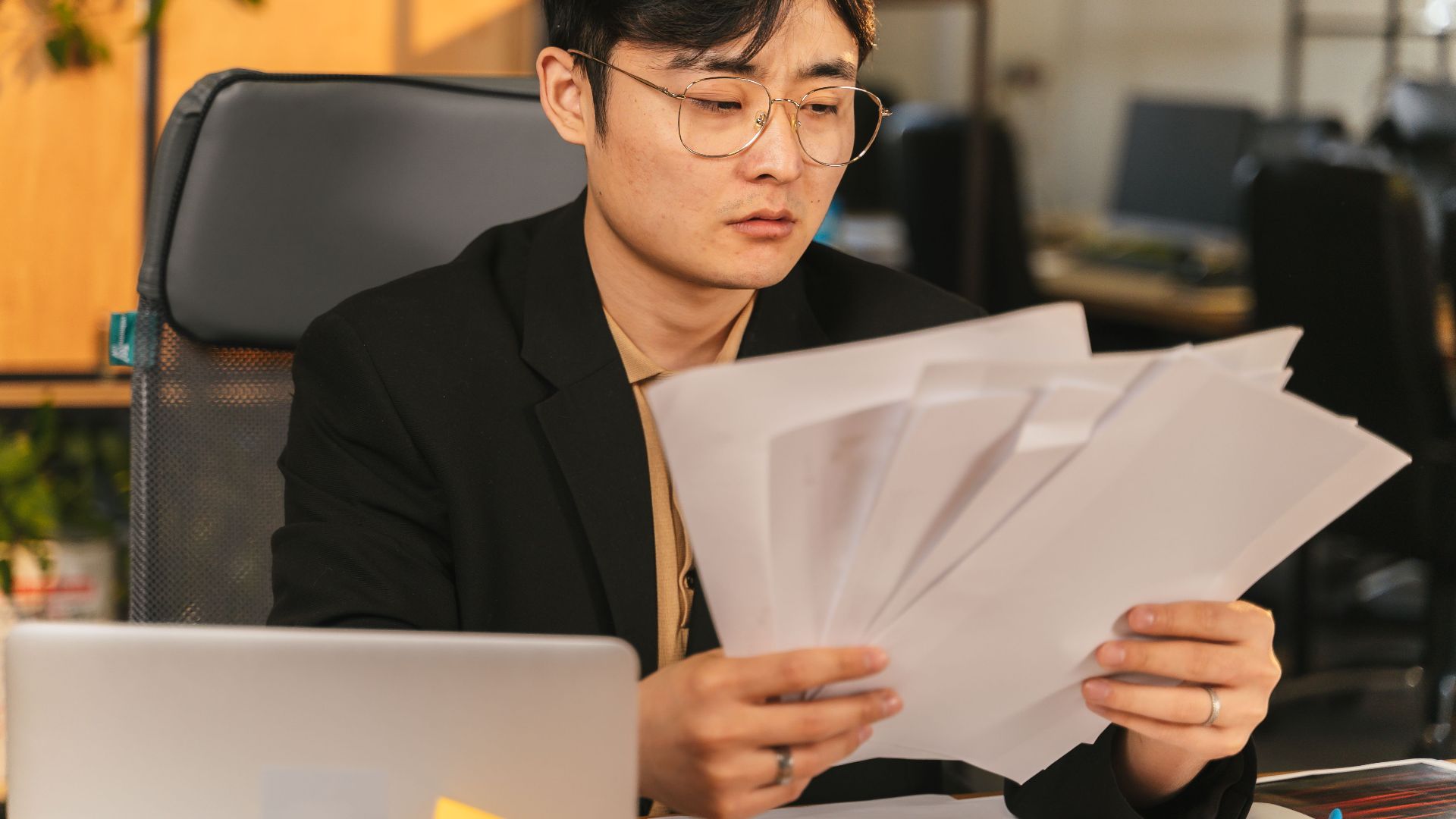 Asian man in glasses and suit analyzing papers at his desk in a modern office.