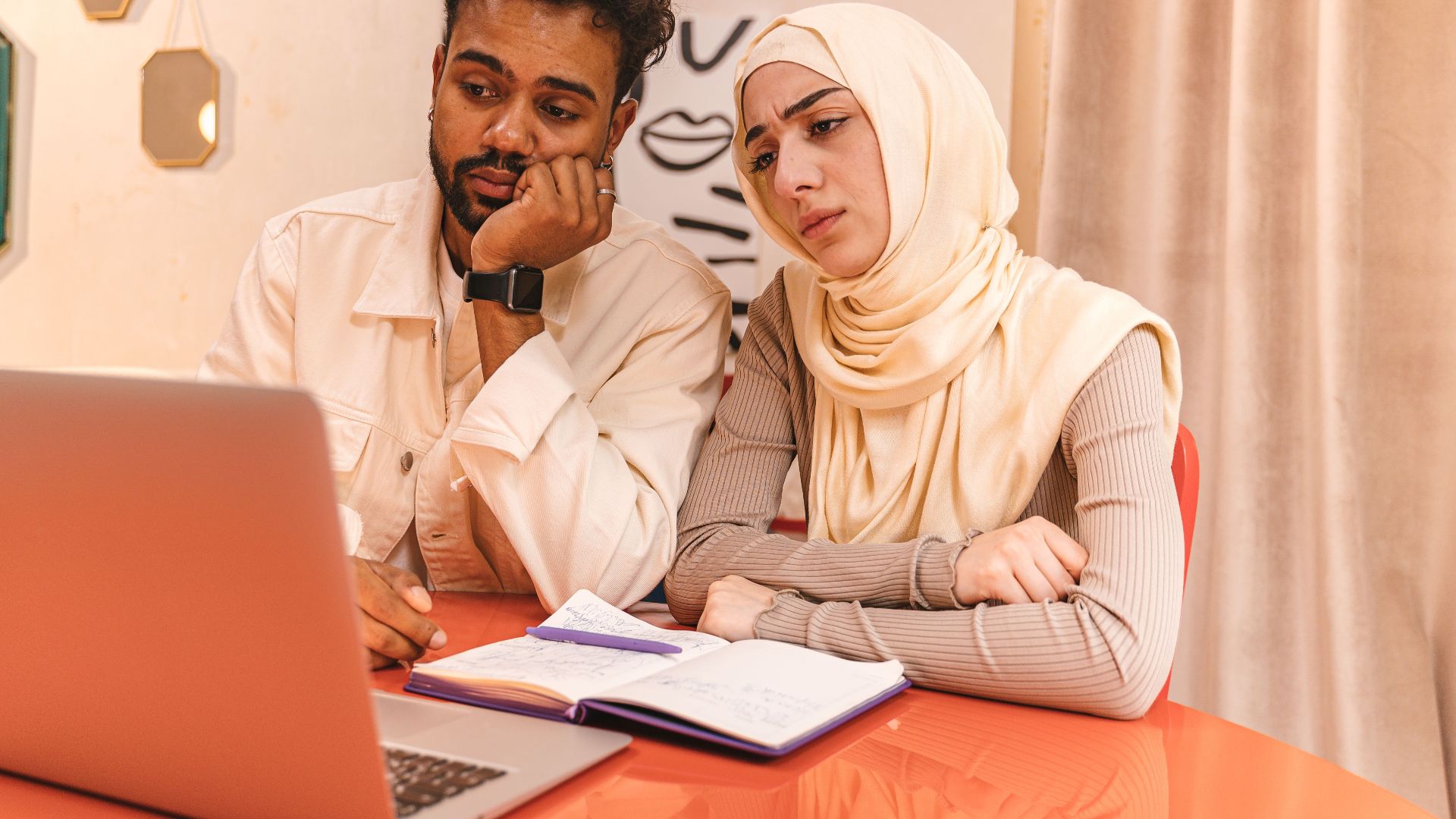 A man and woman in a cozy room focus on a laptop for a collaborative project, surrounded by warm decor.