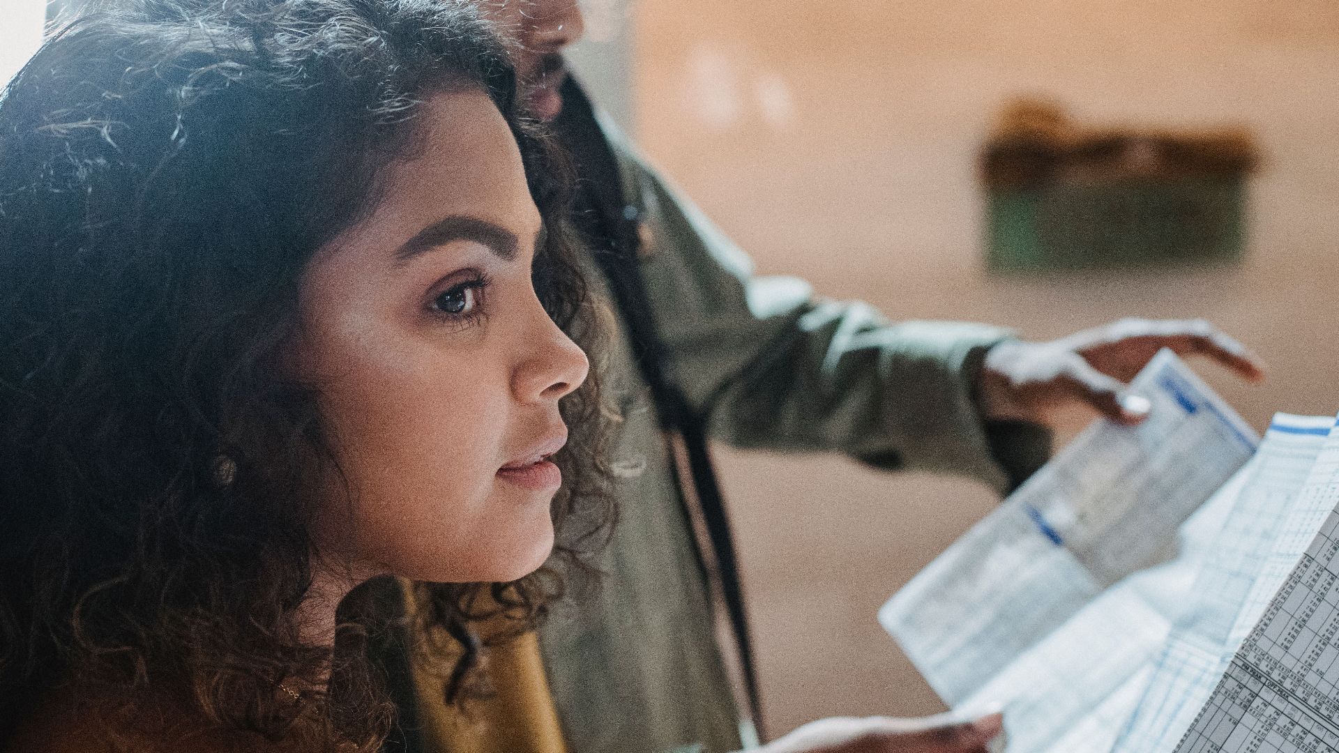 Side view of thoughtful Hispanic woman with dark curly hair standing with black friend reading long paper with tables