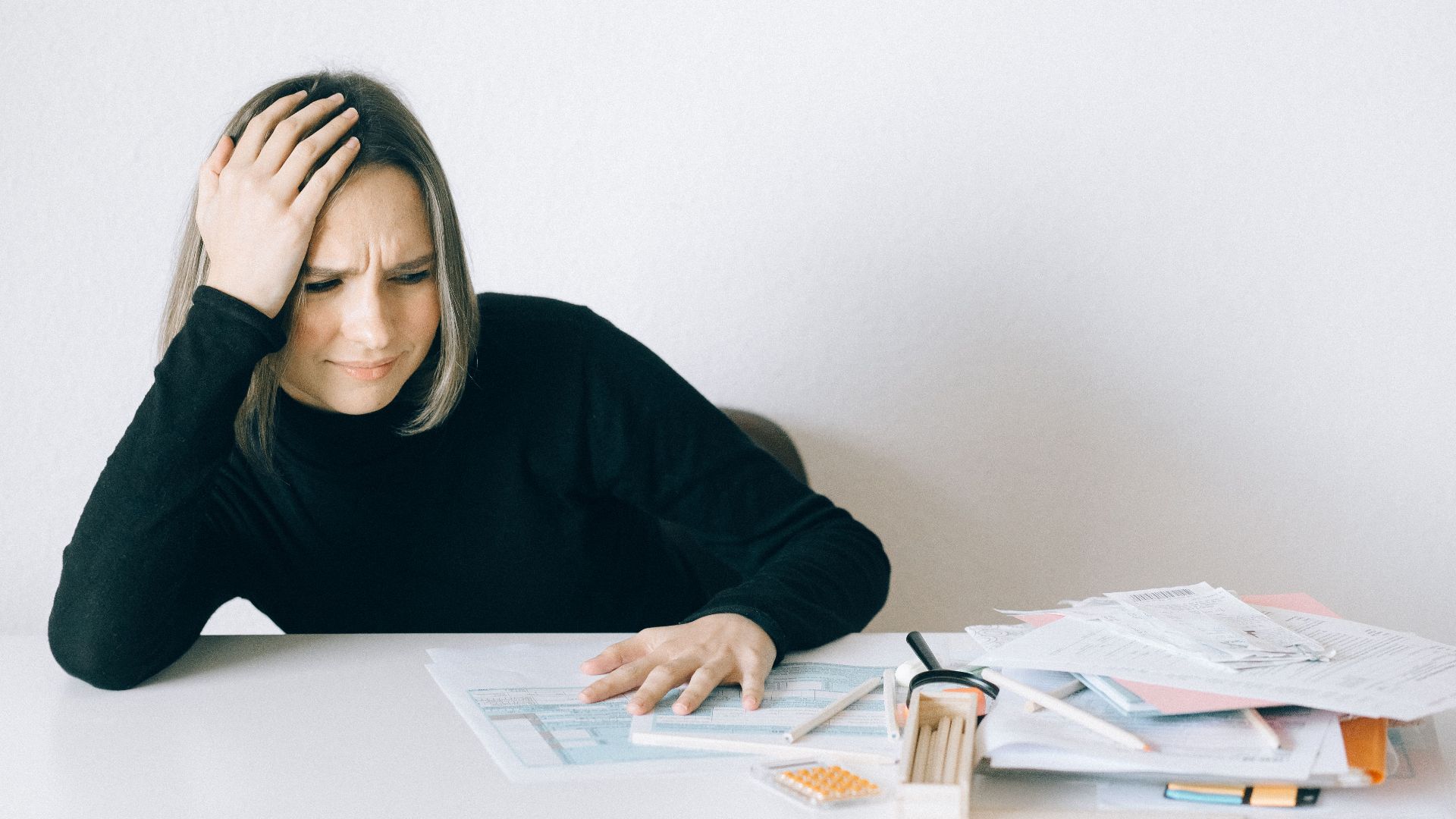 Woman in black sweater stressed with financial paperwork, overwhelmed at white table.