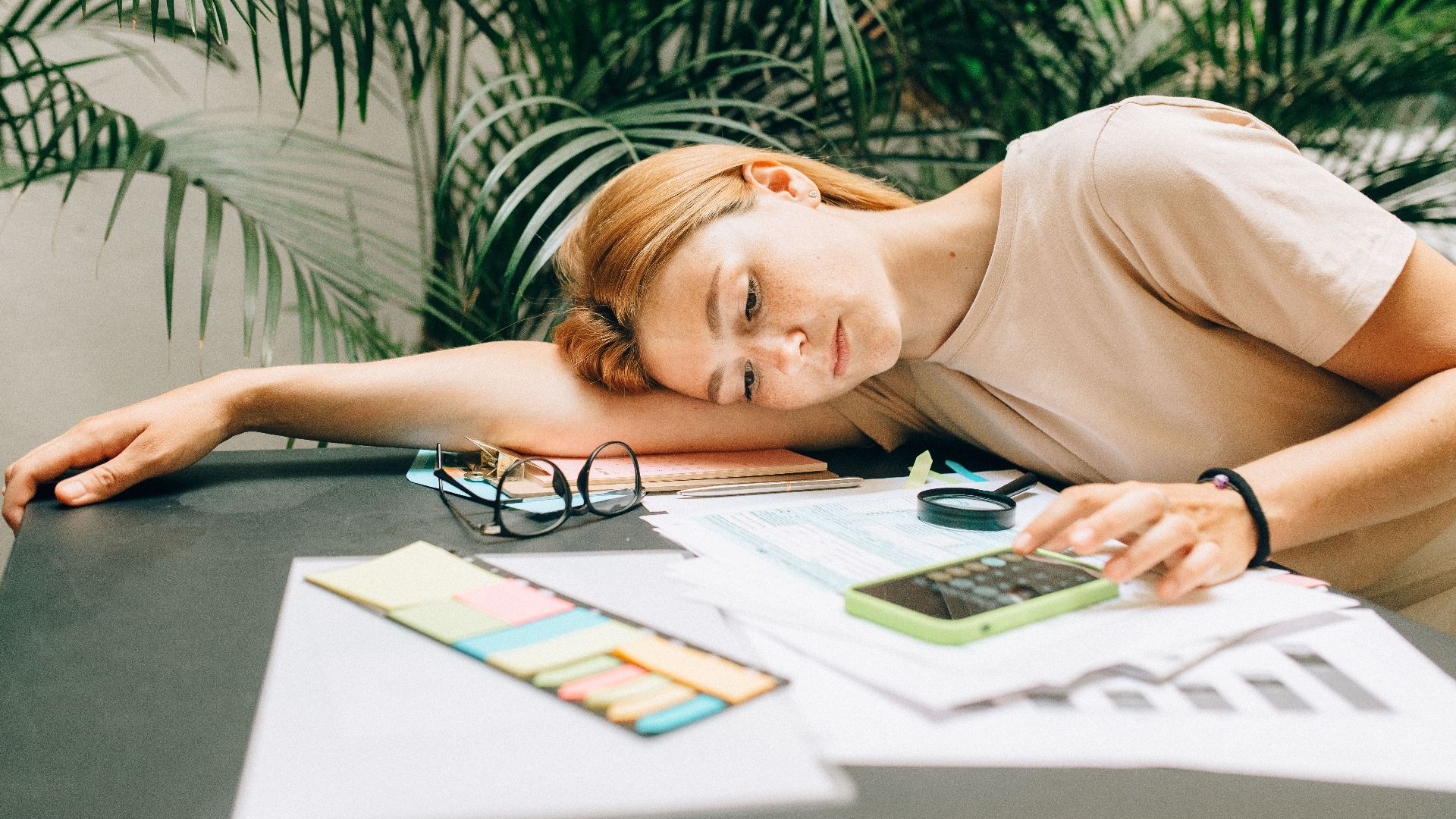 A tired woman calculating her taxes at a desk covered with papers and a smartphone.