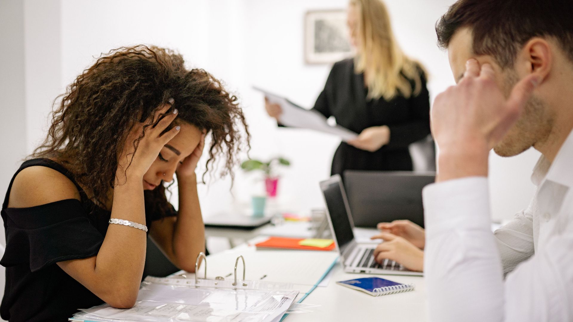 African American woman stressed at office desk with colleagues nearby, conveying workplace pressure.