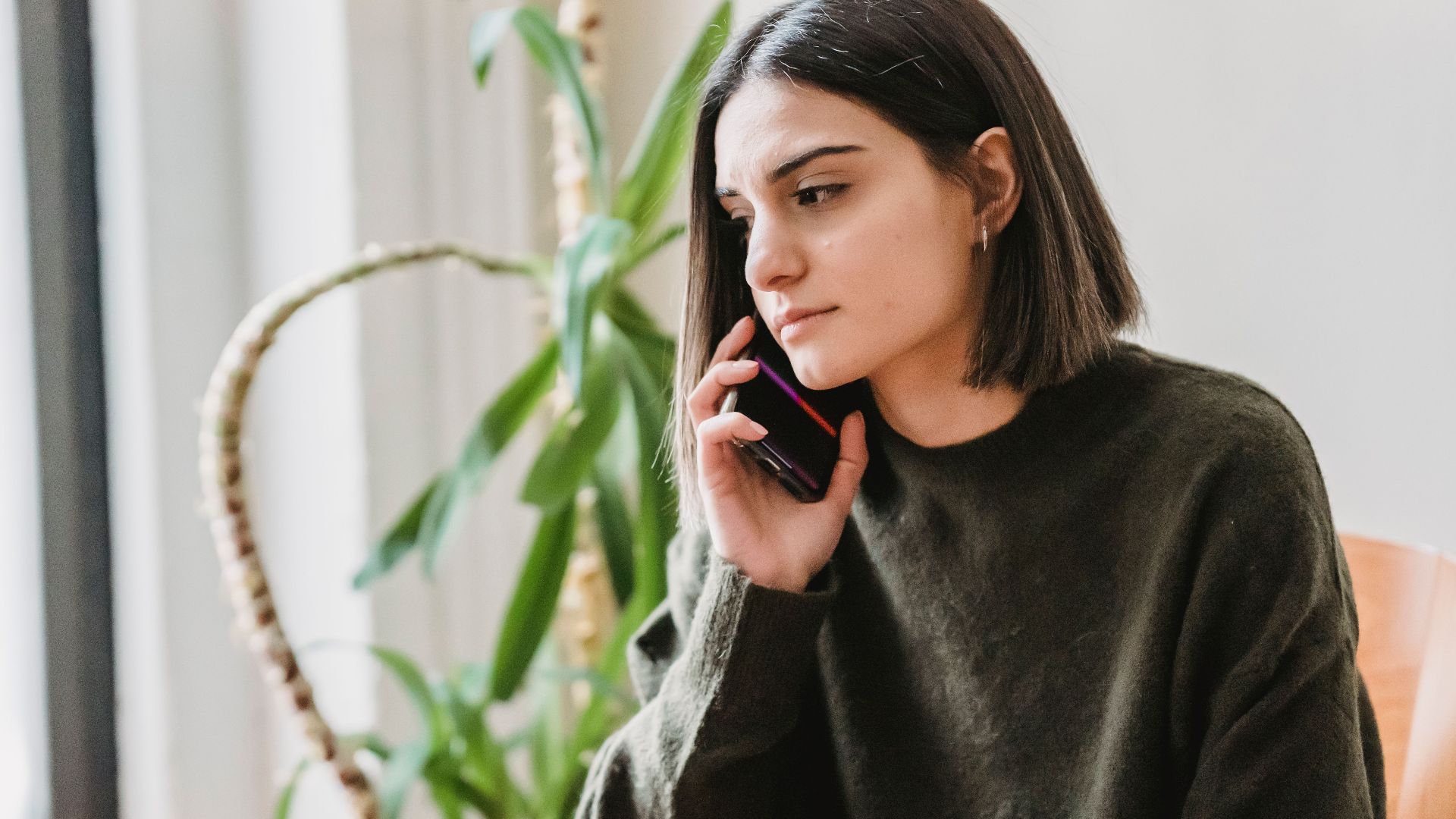 Calm young ethnic female millennial with dark hair in casual clothes having phone conversation while sitting on comfortable armchair at home in daylight