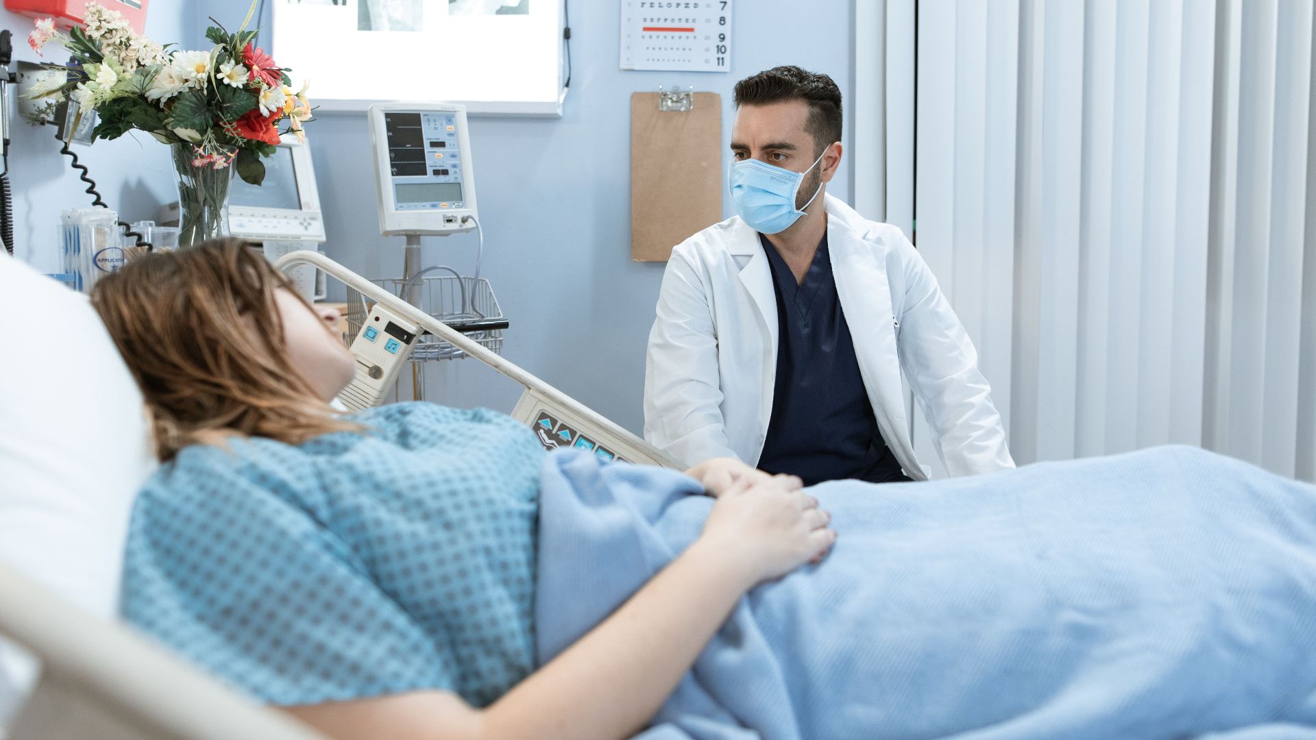 A doctor in a face mask talks to a patient in a hospital bed, providing care and consultation.