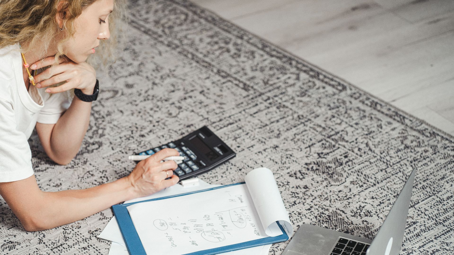 A woman calculates finances at home using a laptop, calculator, and documents on a rug.