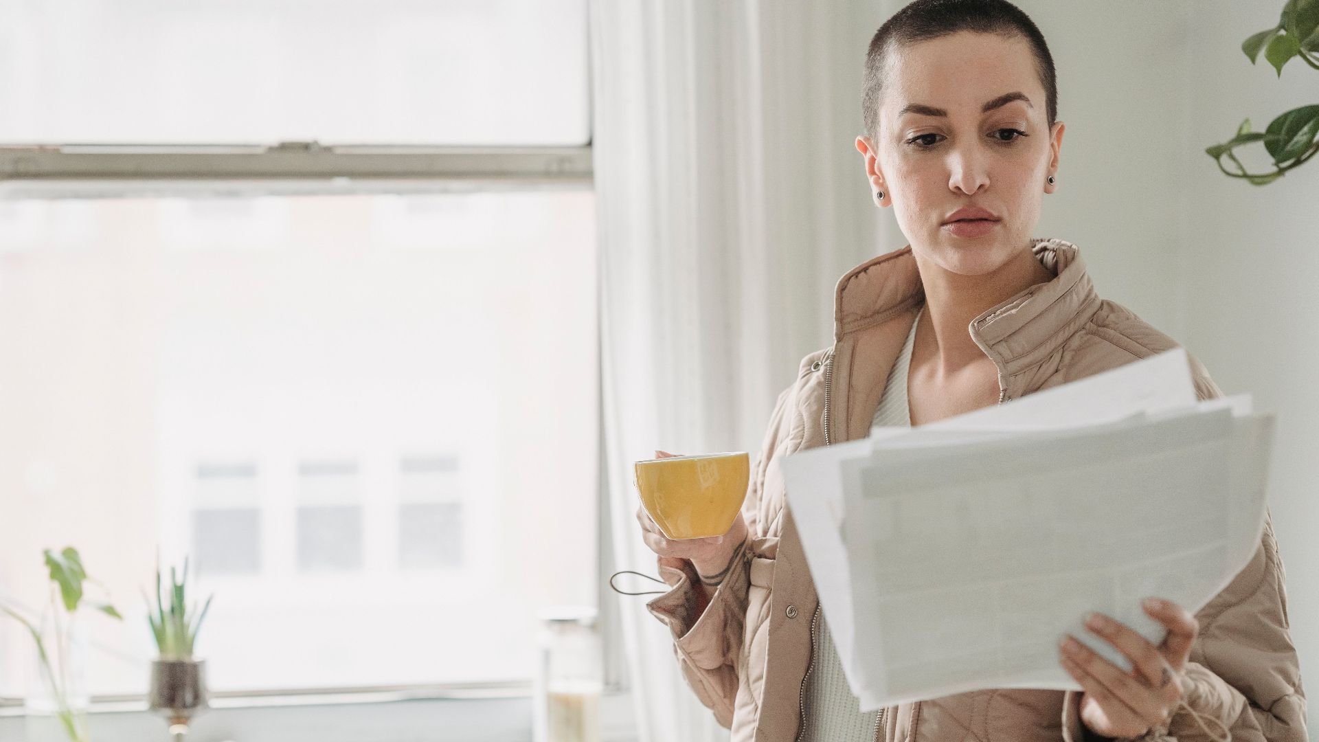 A woman stands by a window holding papers and a coffee cup, in daylight.