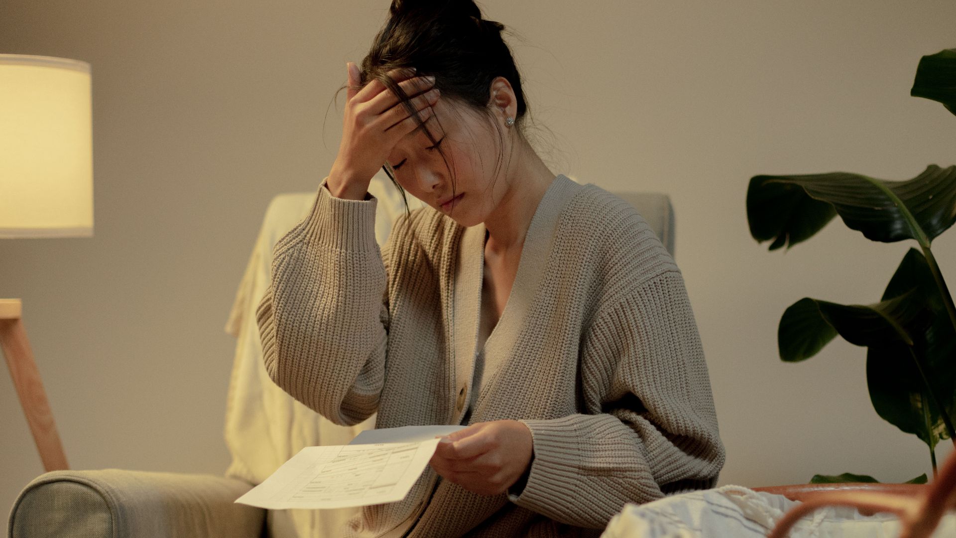A woman sits in a cozy room, stressed while reading paperwork, hand on head.