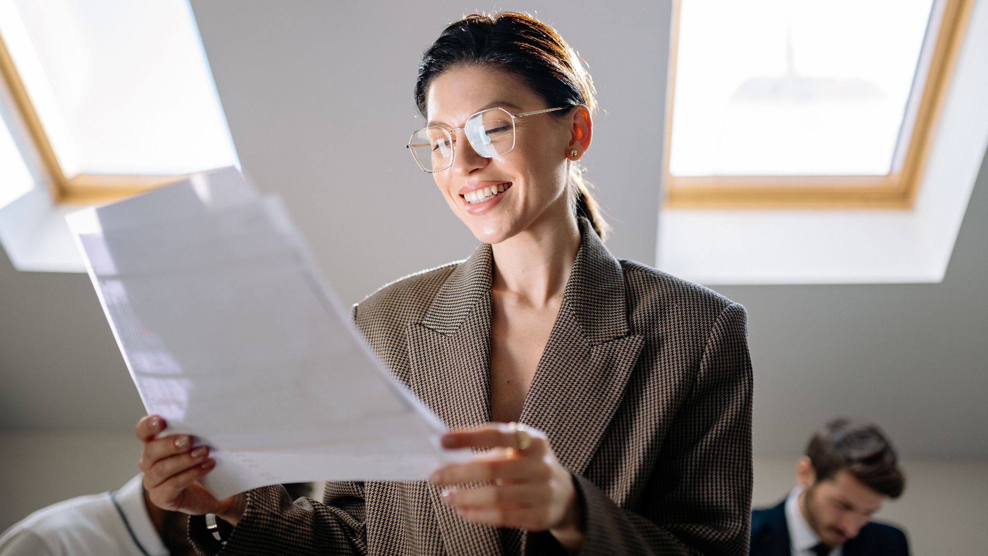 Smiling woman wearing eyeglasses examining paperwork in modern office setting.