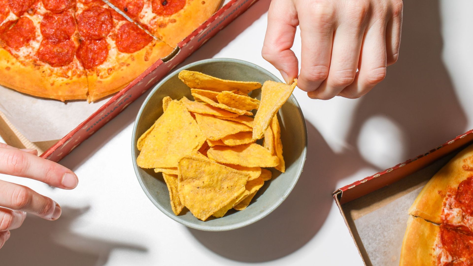 Close-up of pepperoni pizza boxes with a bowl of nachos and hands reaching in, on a bright white table.