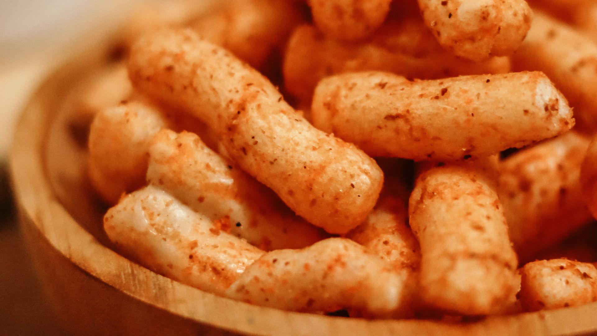A close-up shot of crunchy, spicy snack sticks in a wooden bowl on a table.
