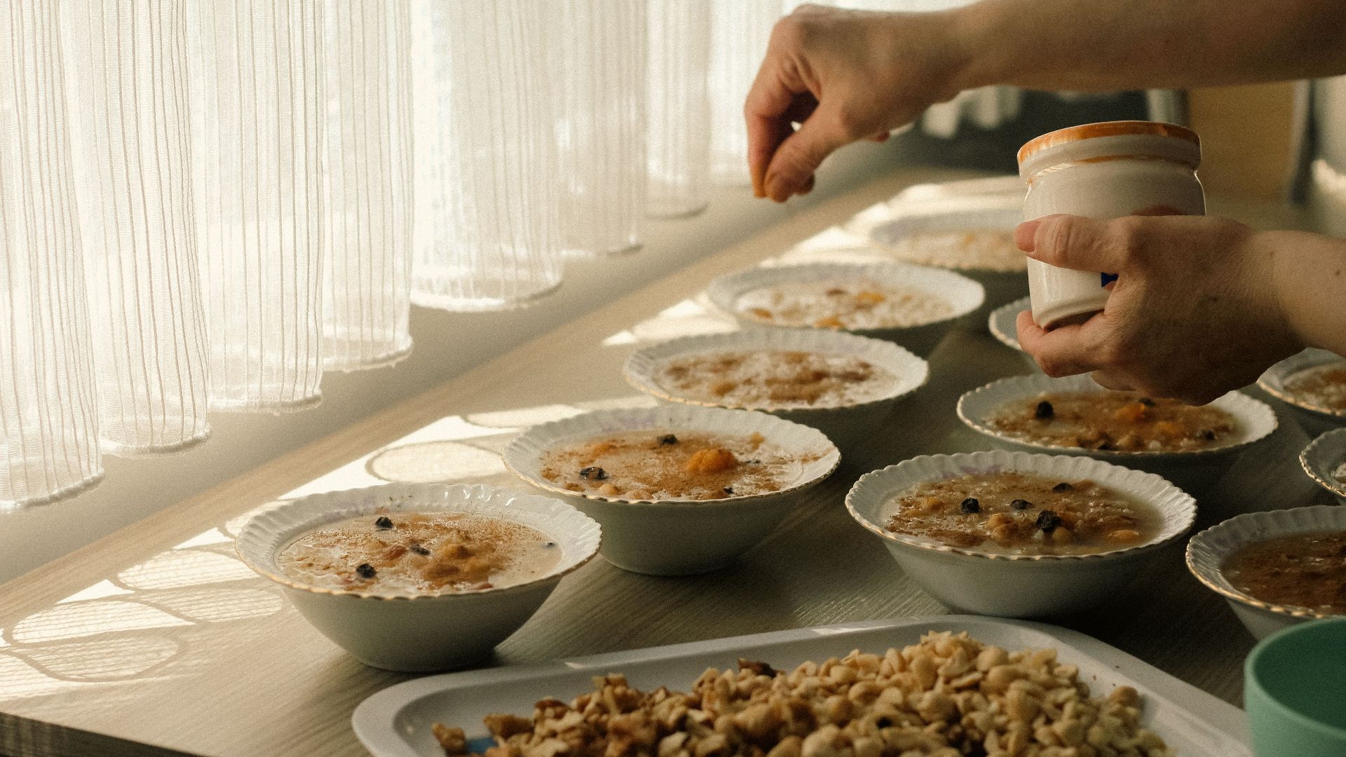 Person adding toppings to bowls of oatmeal on a sunlit table, featuring nuts and raisins.