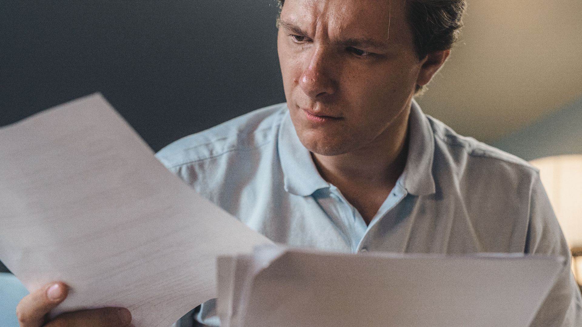 A man intently examines papers, seated indoors under warm lighting, focusing on his work.