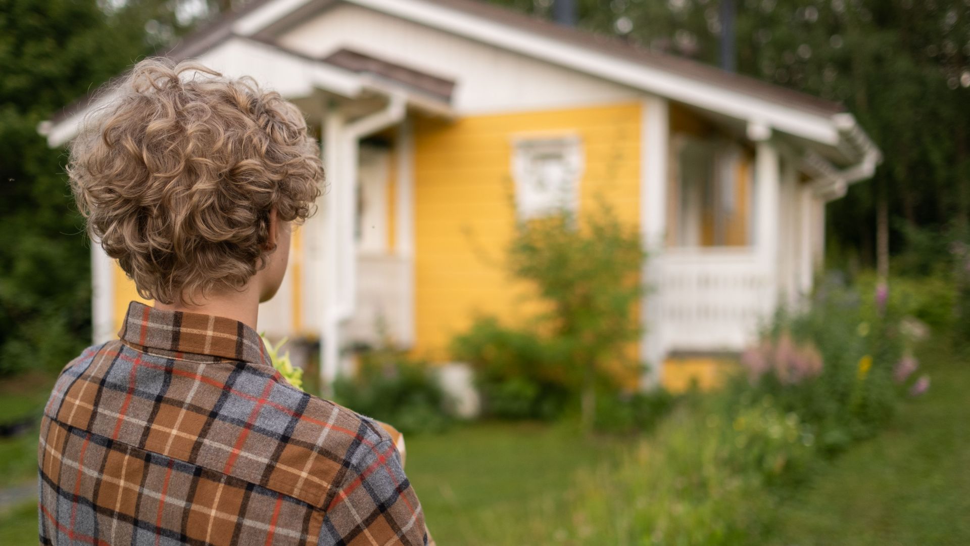 Teen with curly hair in plaid shirt stands facing a charming yellow house amidst lush greenery.