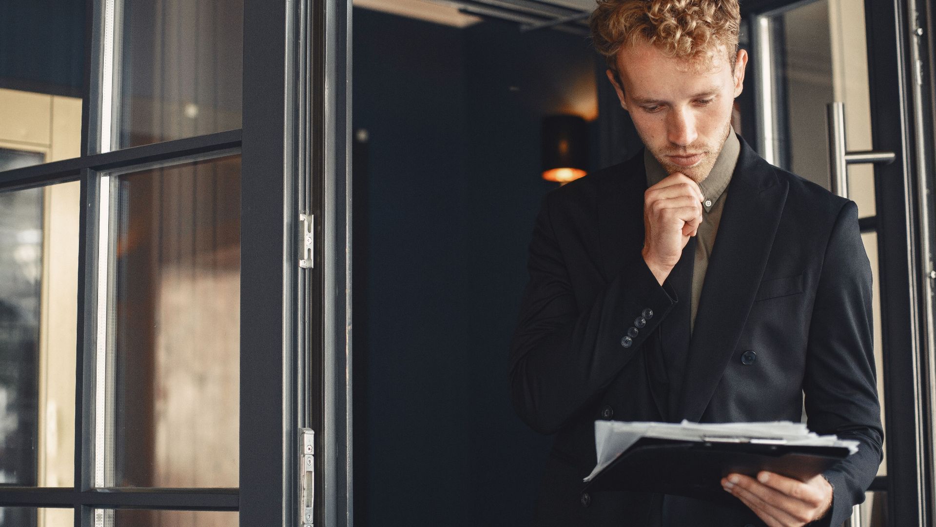 A businessman in a suit thoughtfully reviews documents while standing by a door outdoors.