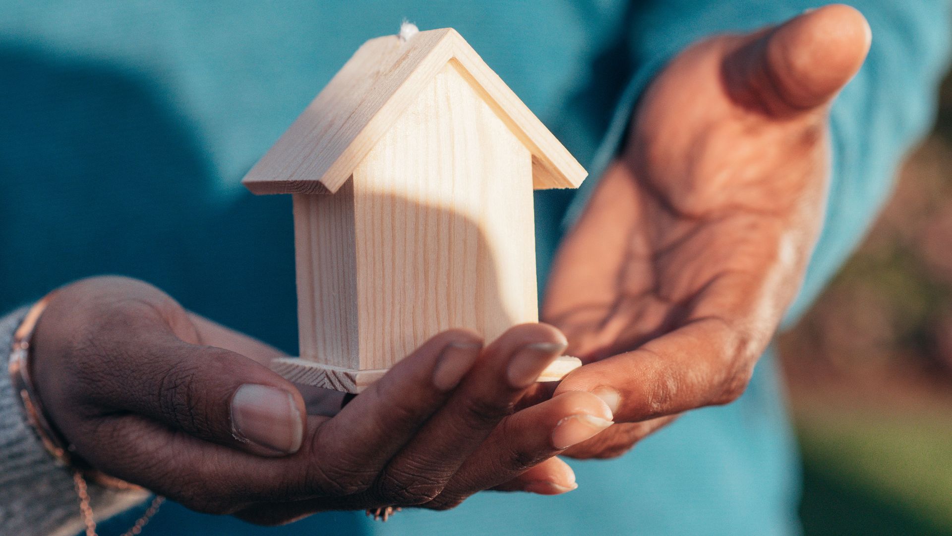 Close-up of hands holding a small wooden house model outdoors, symbolizing real estate investment.