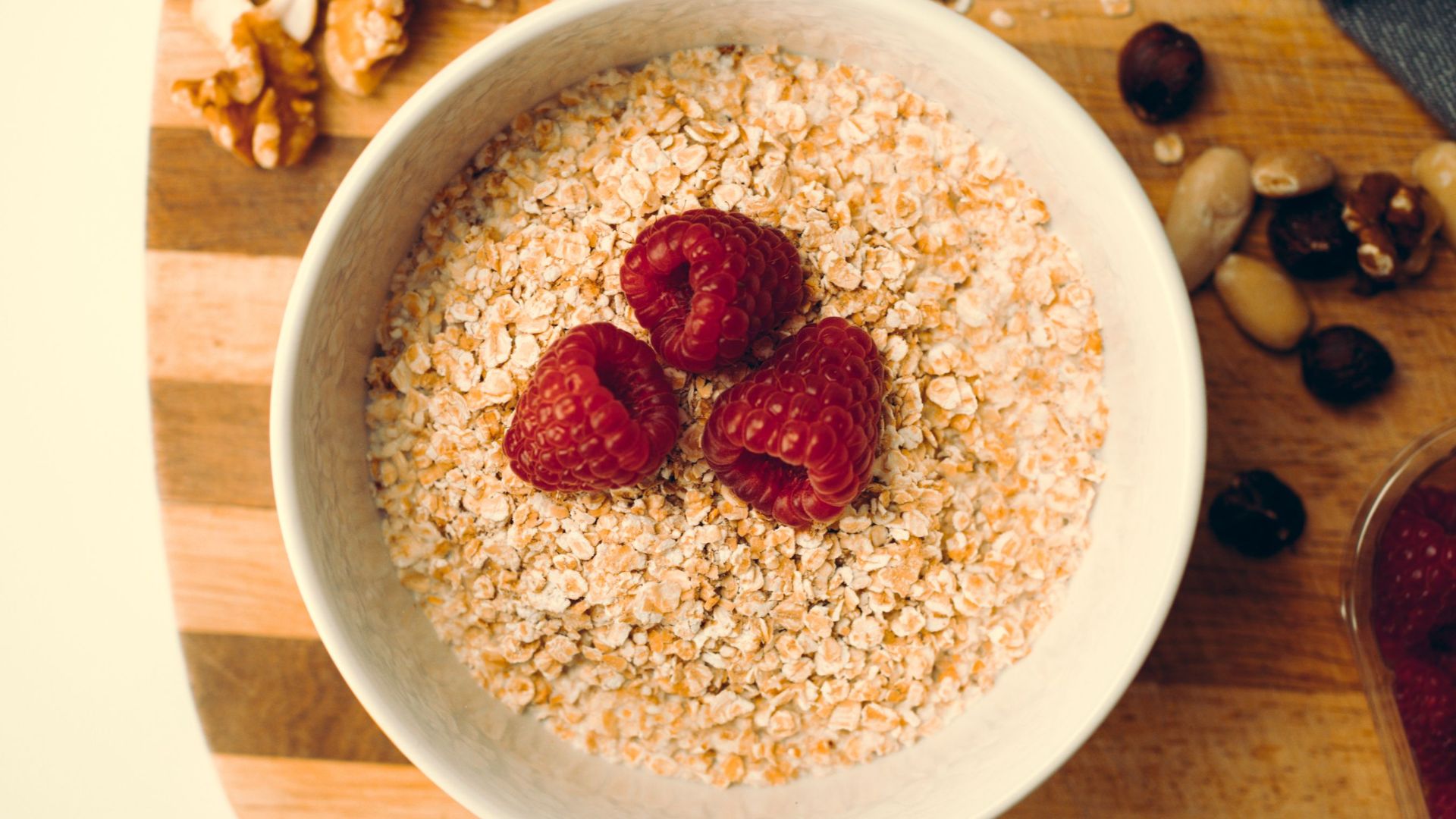 Top view of a white bowl with oatmeal, raspberries, and nuts on a wooden board, perfect for a healthy breakfast.