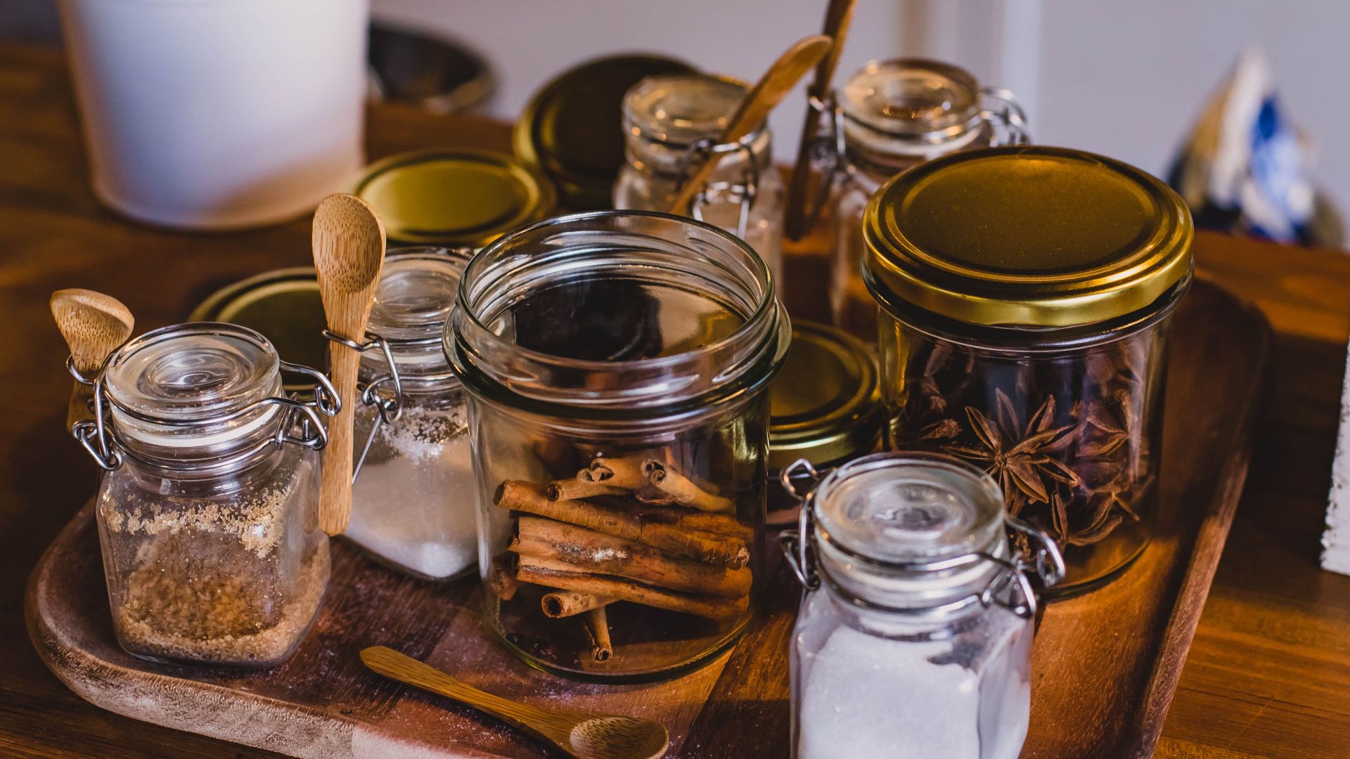 An arrangement of spice jars and cinnamon sticks on a wooden tray in a cozy kitchen setting.