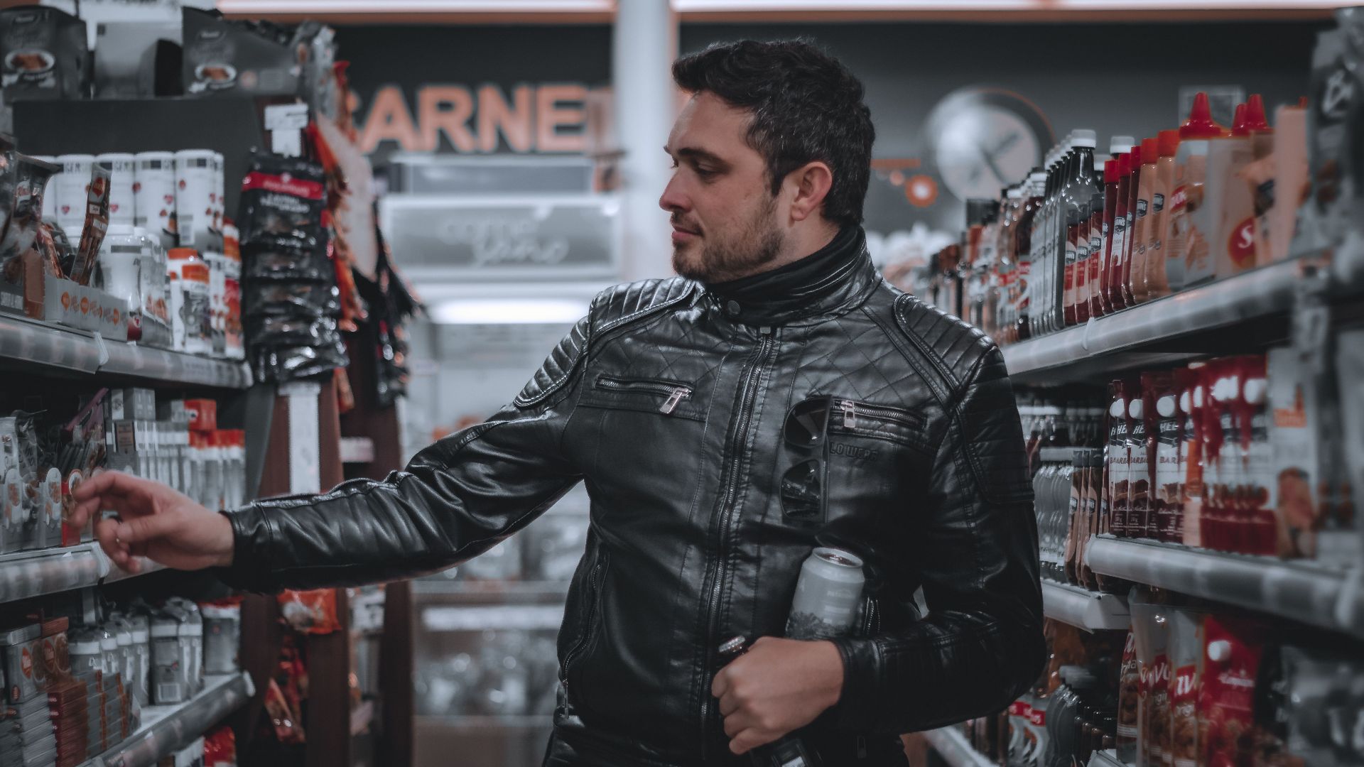 Stylish man browsing products in grocery store aisle, wearing a black leather jacket and holding a drink.