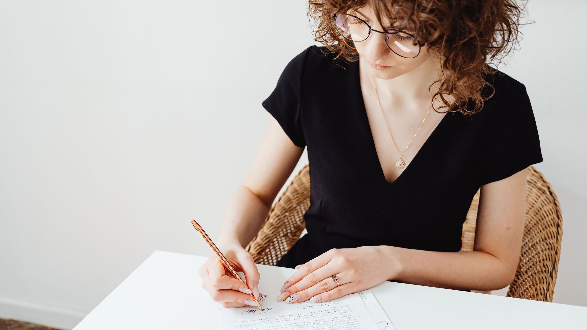 A woman in glasses and black top writing on documents at a white table, focusing intently.