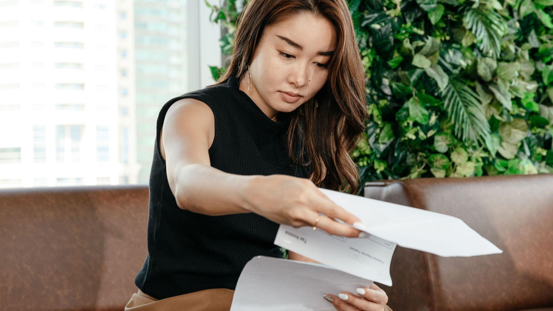 Focused Asian female turning pages of document while sitting on sofa during paperwork in modern workspace with green deciduous plant