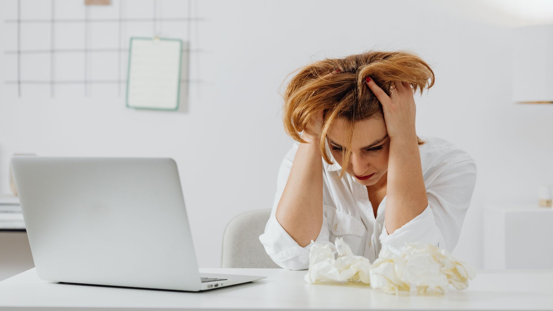 Frustrated woman in white shirt at desk with laptop, feeling overwhelmed.